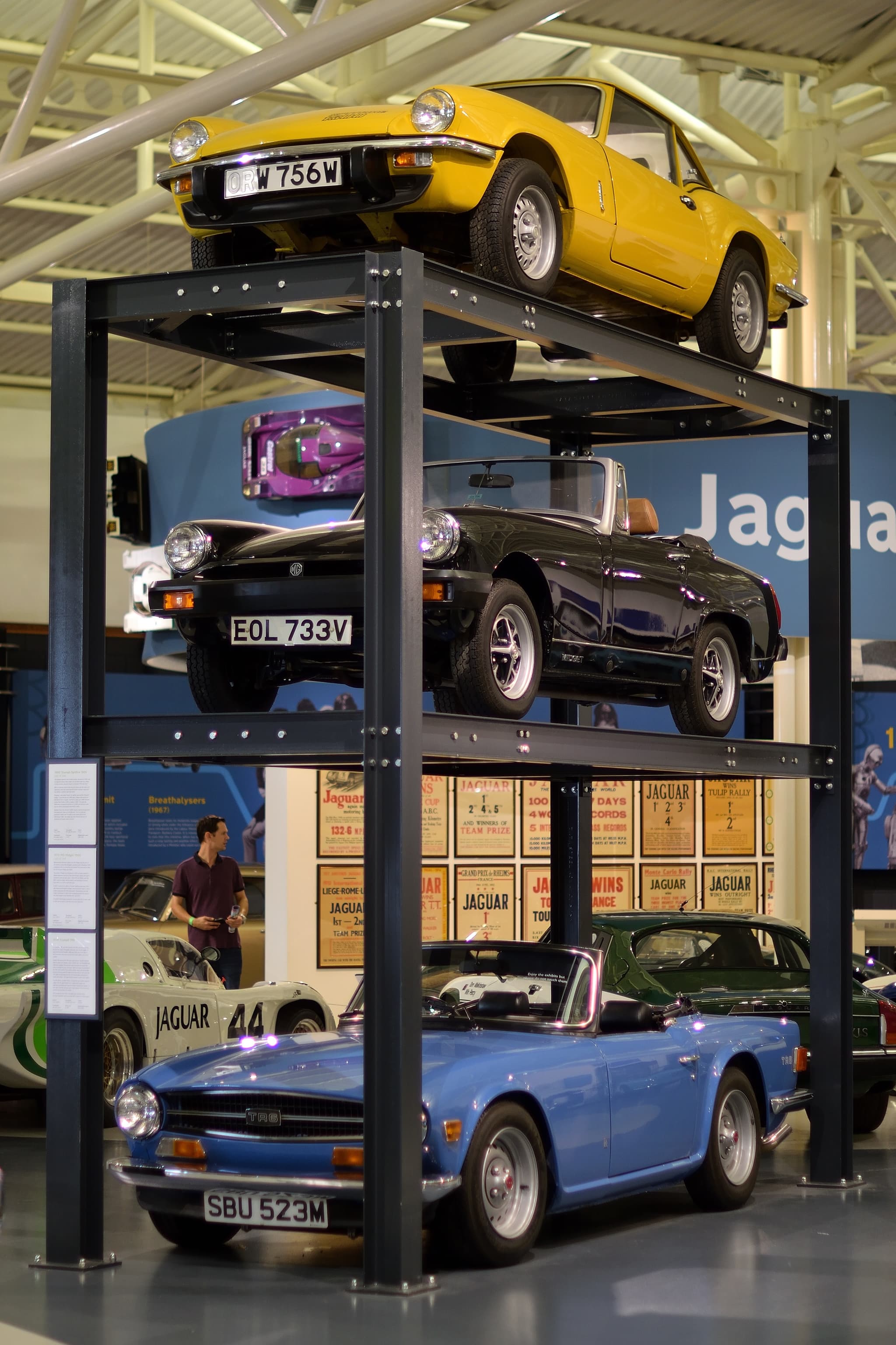 A yellow Spitfire with a hardtop on the top tier of a three-level car display rack in a museum. Opens a larger view