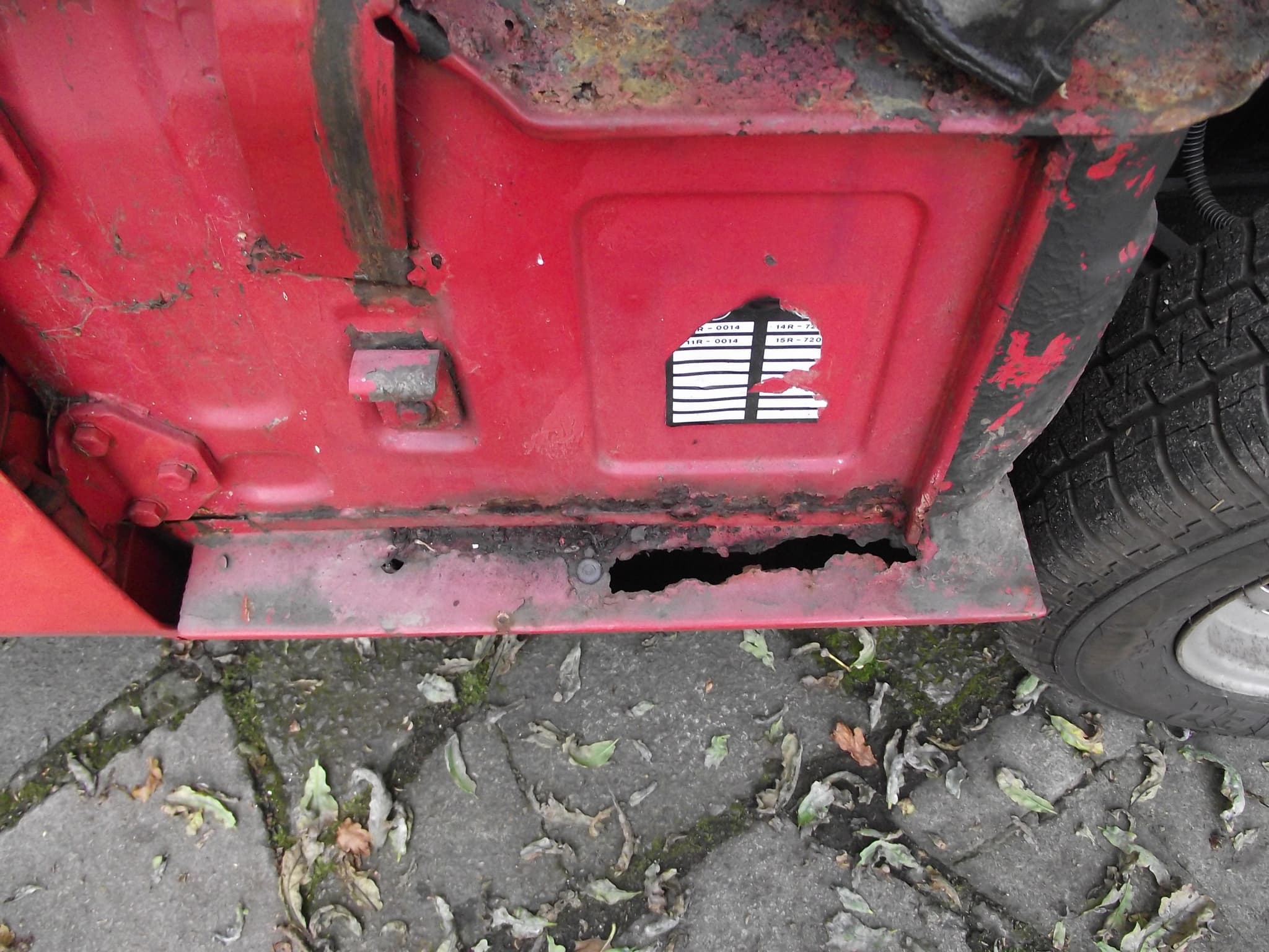 The side of a red bulkhead above the front end of the sill, the outer sill rusted through into a ragged hole above paving.