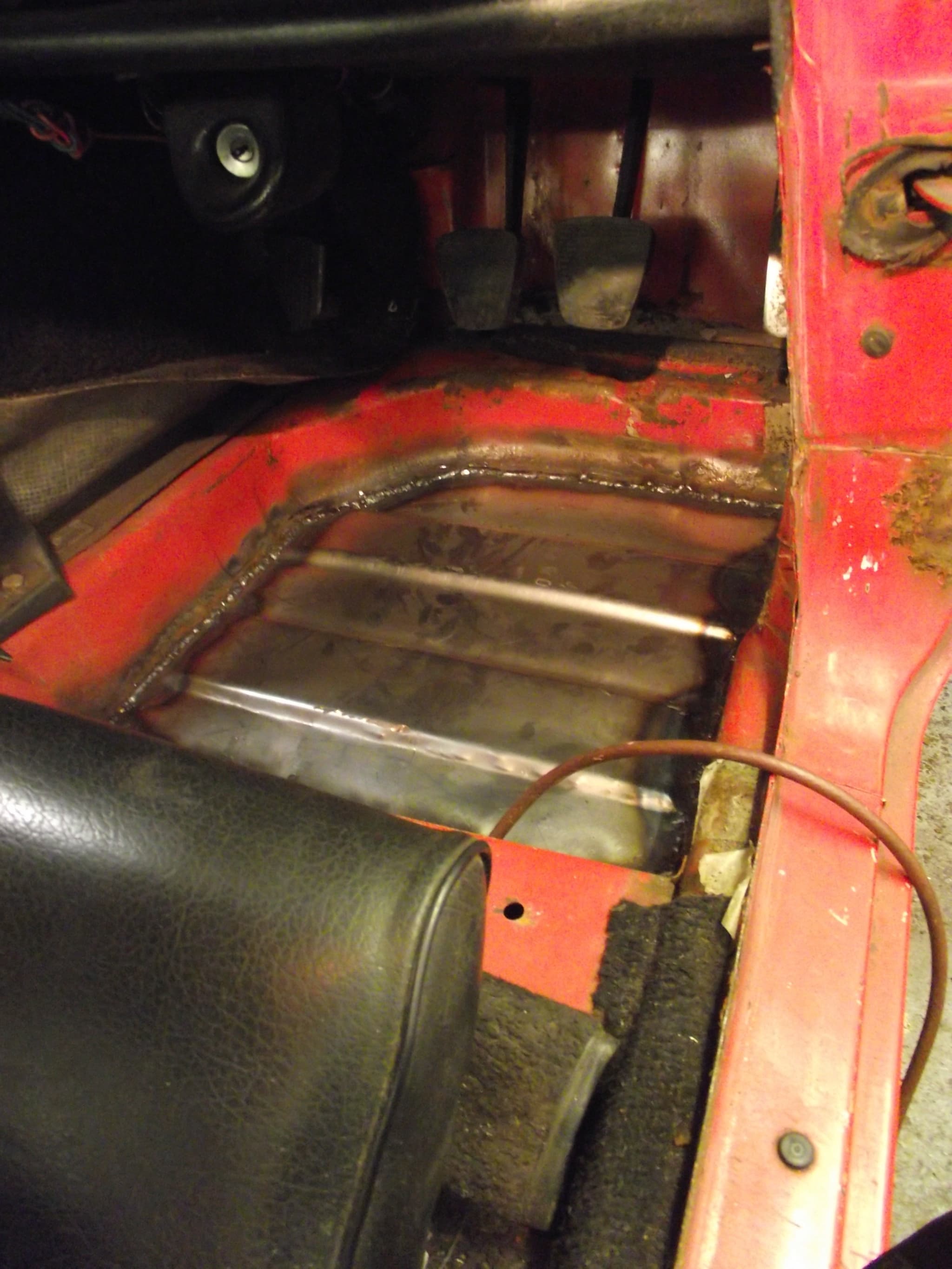 A car's driver's footwell from above, a bare steel floor panel welded in below the pedals, the seat tipped forward.