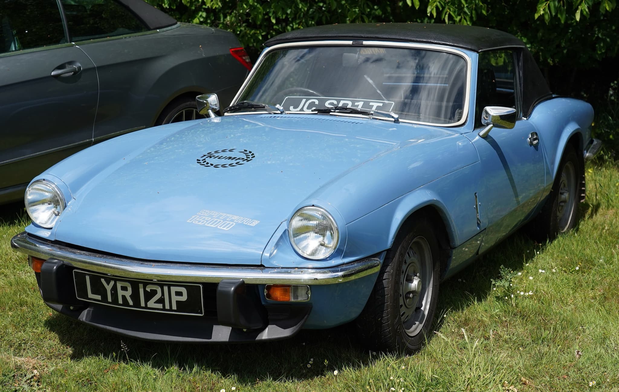 A pale blue Spitfire 1500 with its black hood up, parked on a sunny lawn by a hedge. Opens a larger view