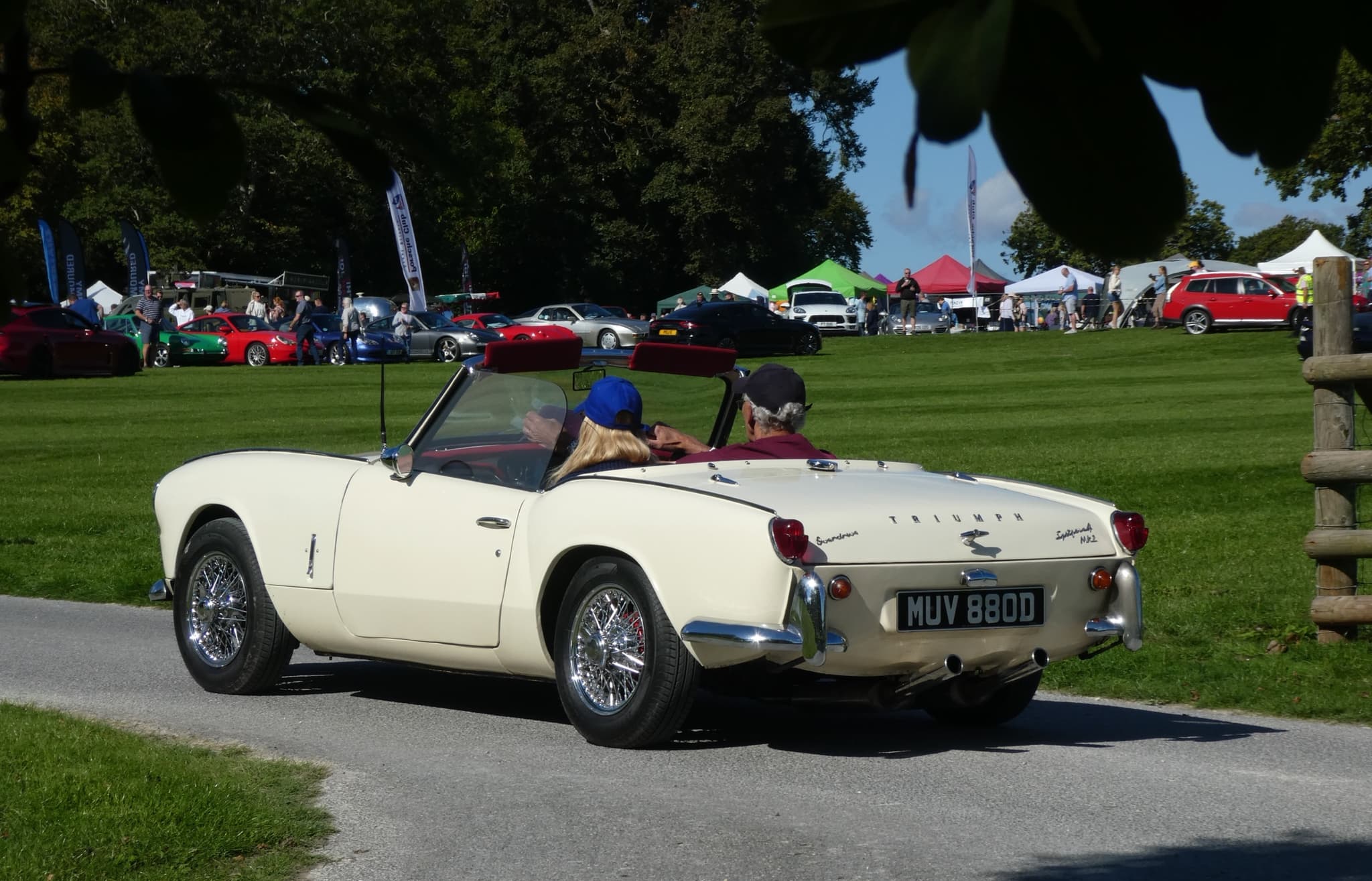 A white Spitfire 4 Mk II with two people aboard driving away along a park drive past a show field.