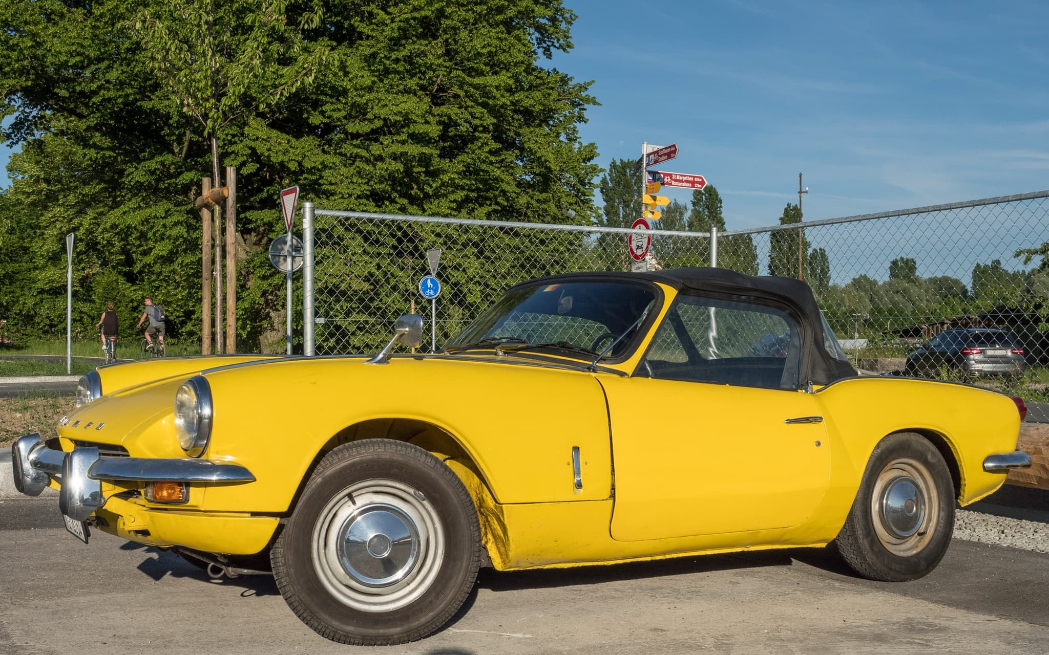 A yellow Spitfire Mk III with its black hood up, parked by a wire fence under trees.