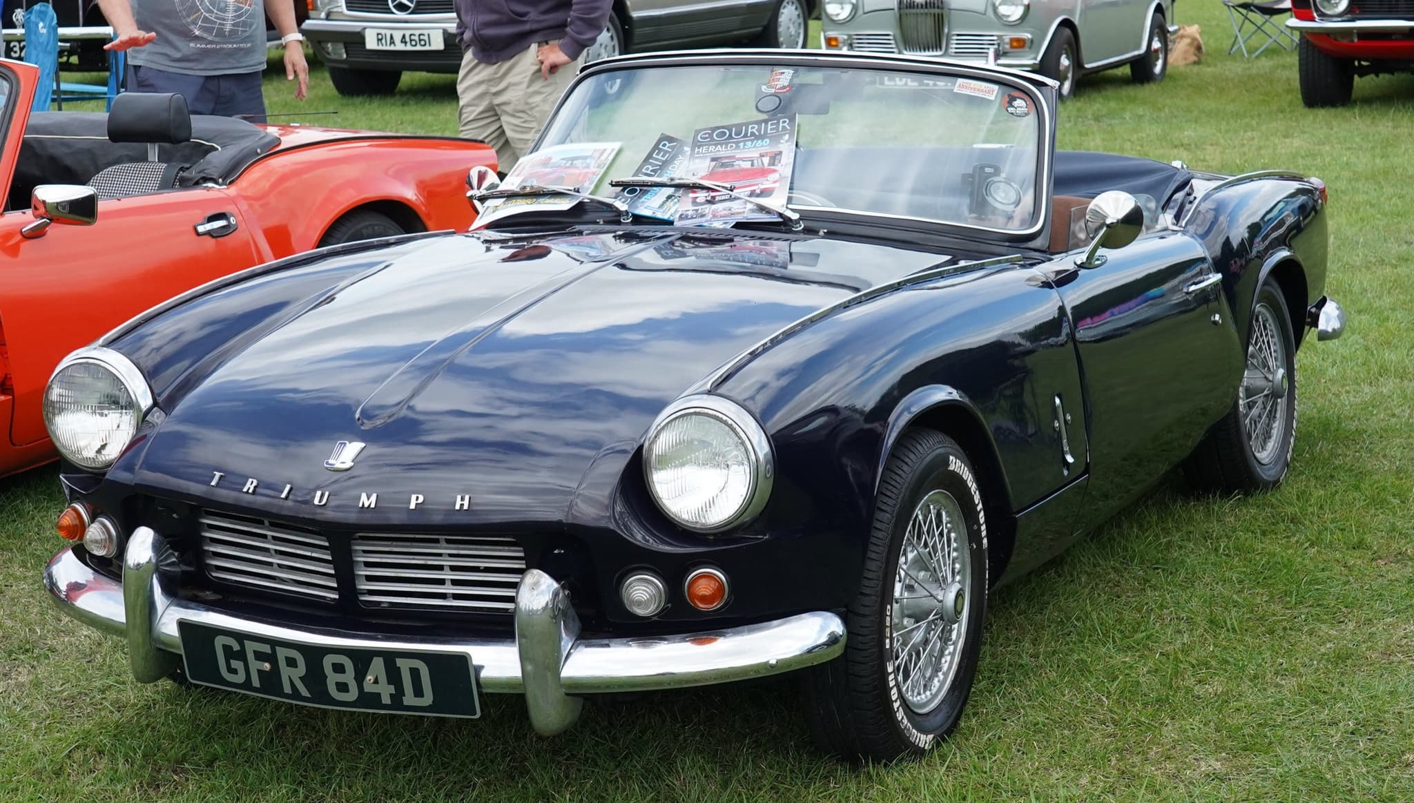A dark blue Spitfire 4 Mk II with wire wheels and its hood stowed, on a grassy show field.