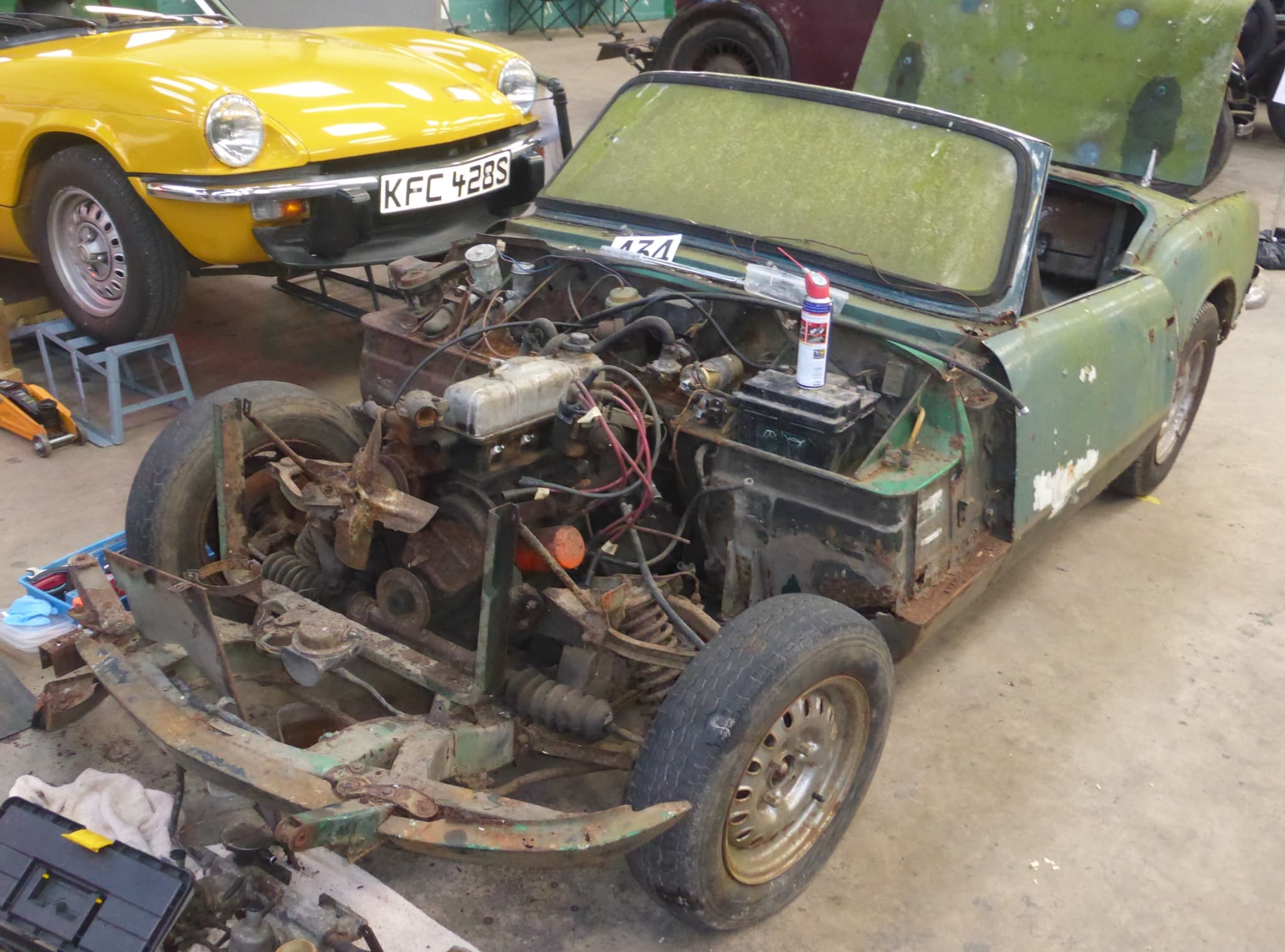A derelict green Spitfire without its front bodywork, showing a rusted chassis, coil springs and the engine, a yellow Spitfire behind.