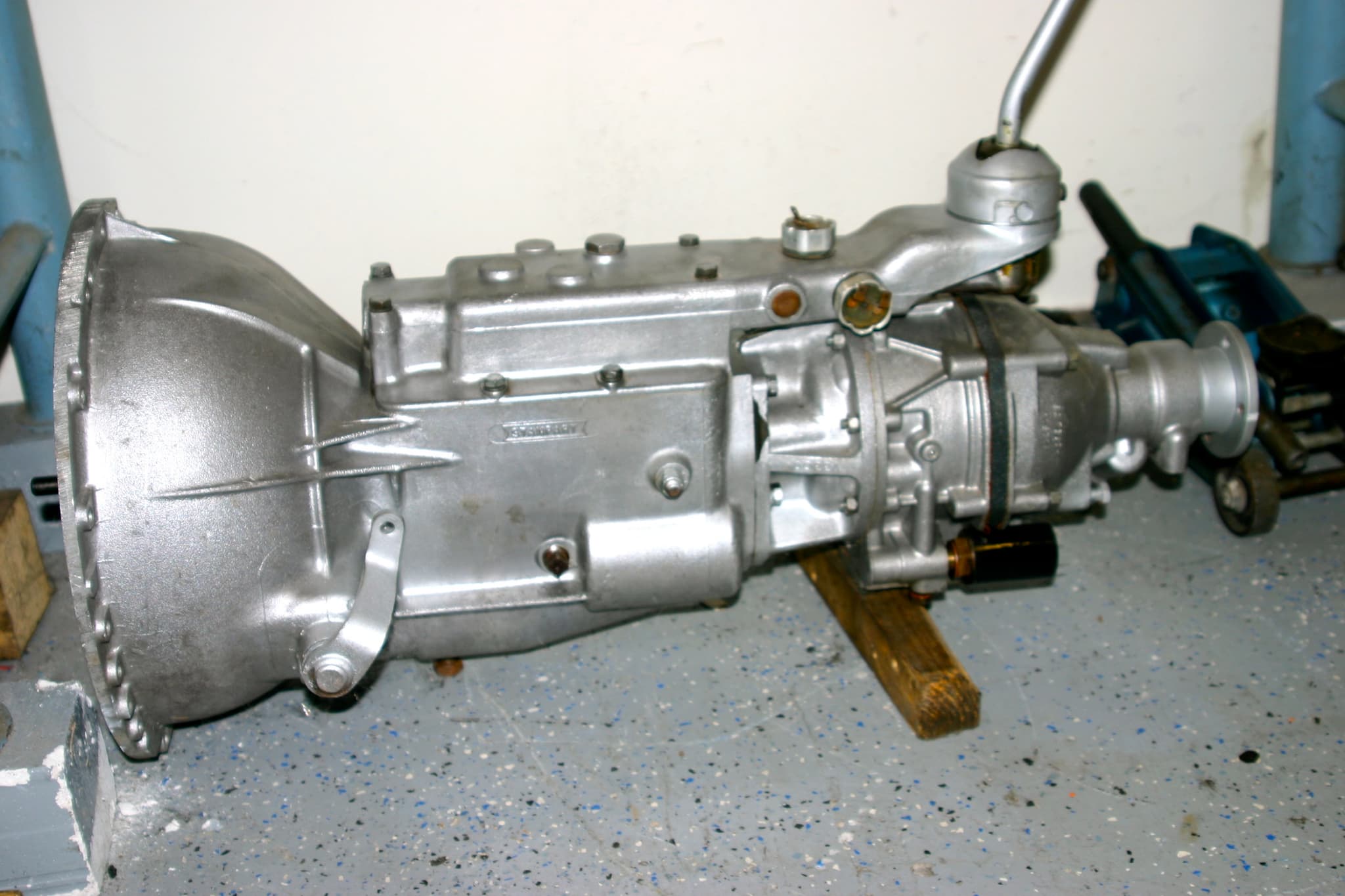 A bare aluminium gearbox with an overdrive unit bolted behind it, its gear lever upright, on a speckled garage floor.
