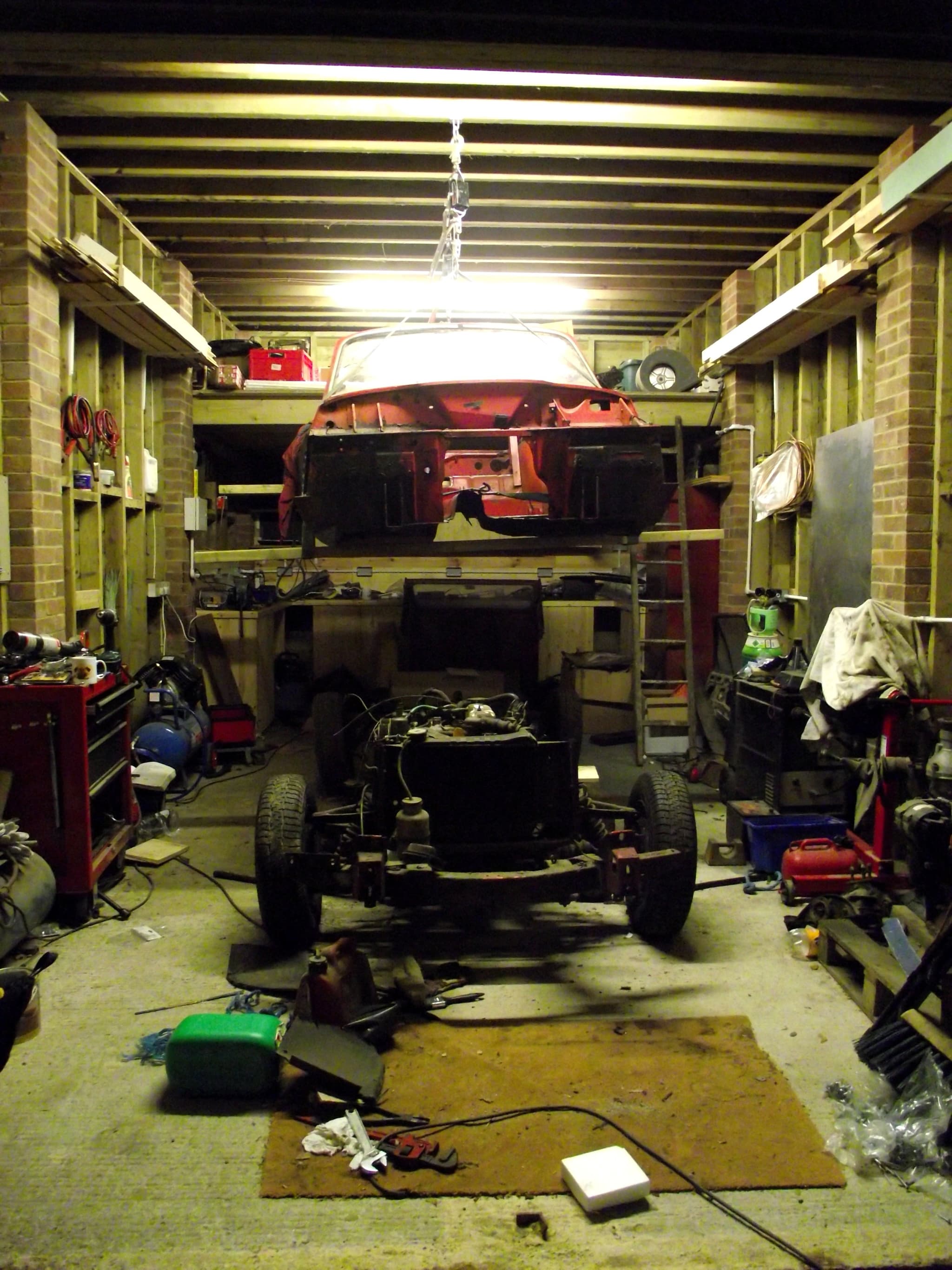 A bare red Spitfire body hoisted to a garage ceiling above its separate chassis and engine.
