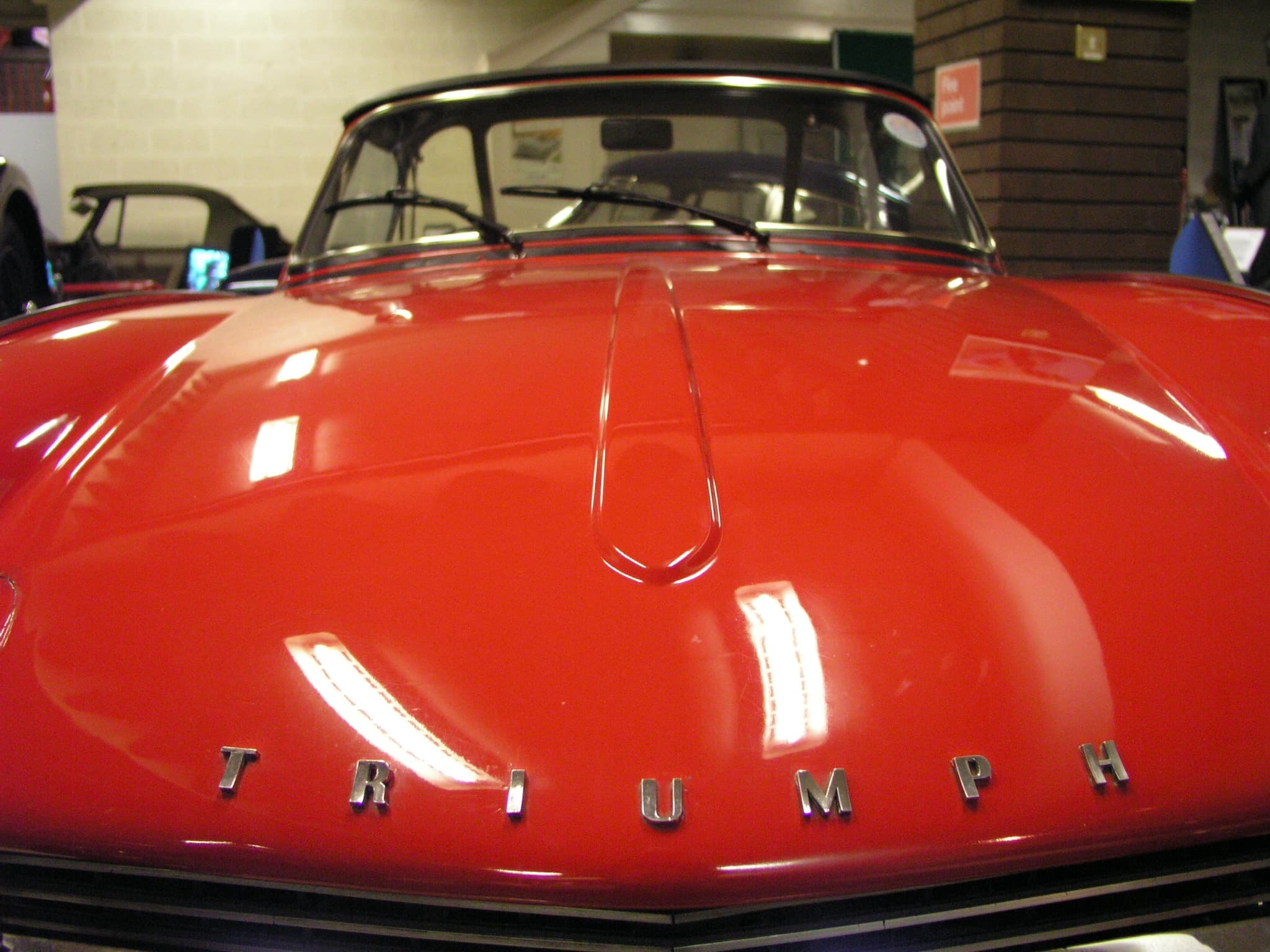 Chrome TRIUMPH letters across the nose of a red Spitfire bonnet, below its teardrop-shaped centre bulge. Opens a larger view