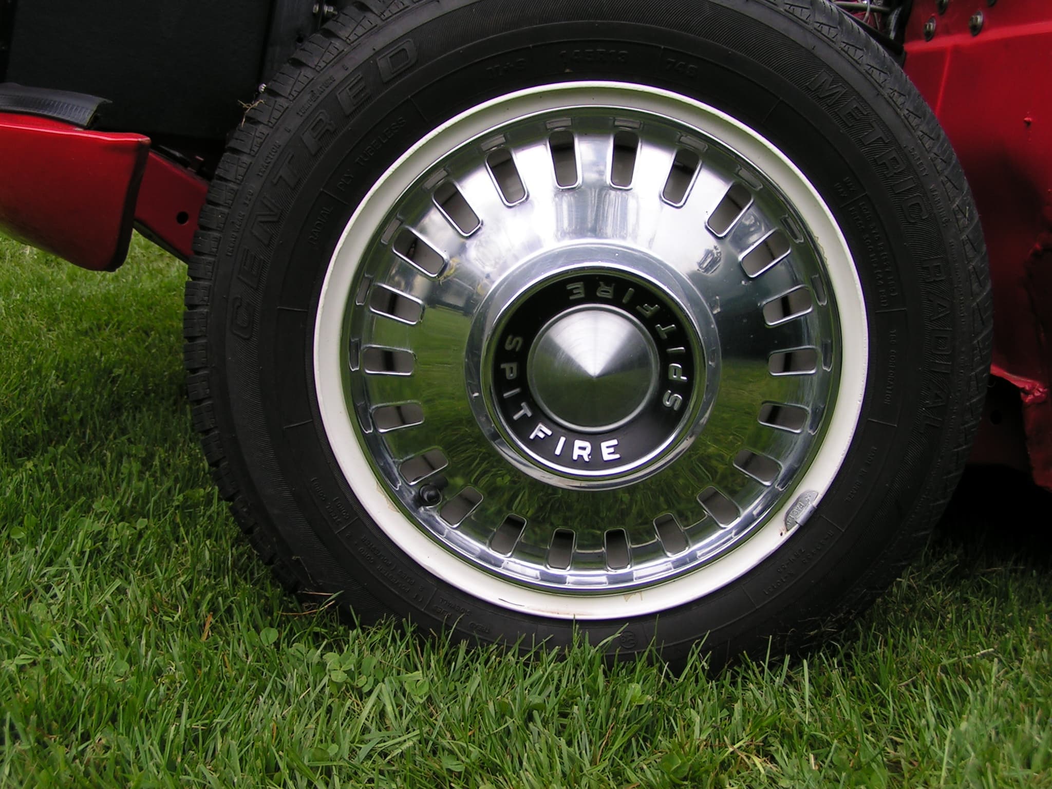 Close-up of a chrome slotted hub cap with a black centre lettered SPITFIRE on a white wheel. Opens a larger view