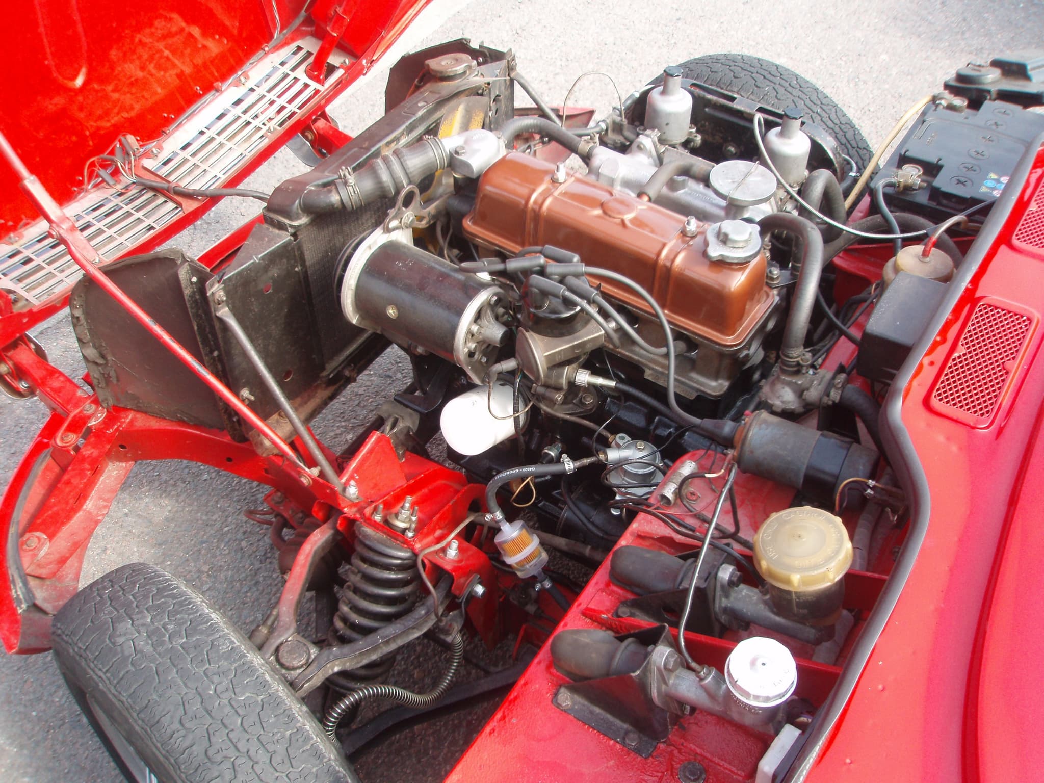 An open Spitfire engine bay with the bonnet tipped forward, showing twin carburettors, a brown rocker cover and front suspension. Opens a larger view