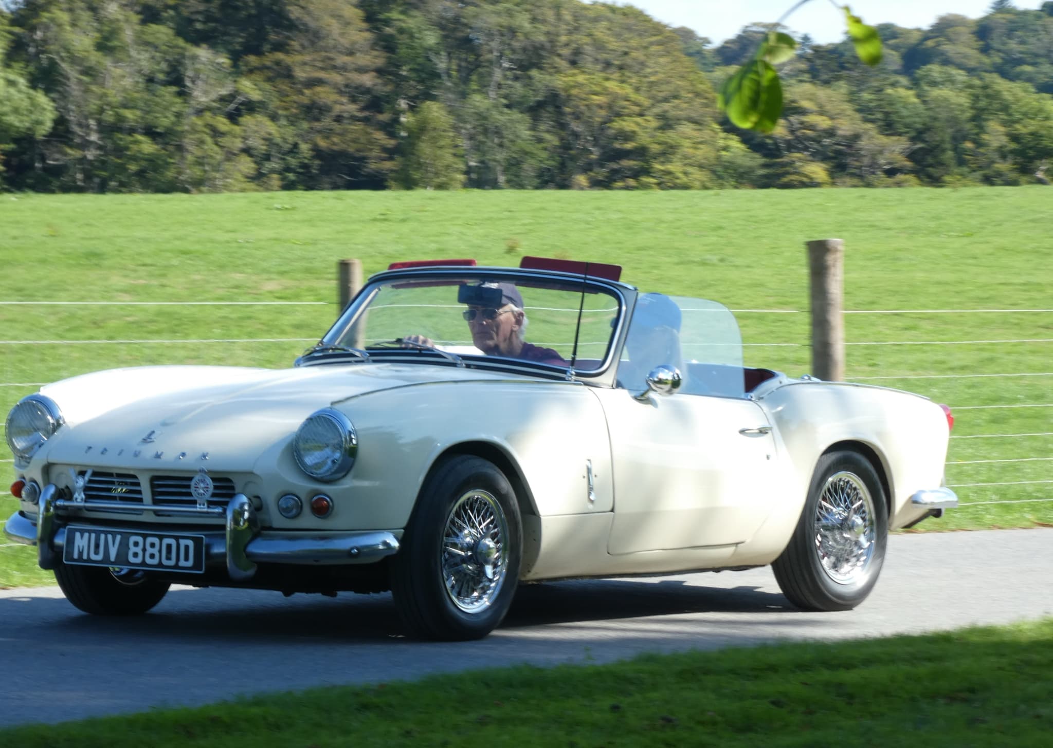 A white Spitfire 4 Mk II on wire wheels driving along a park road past a meadow.
