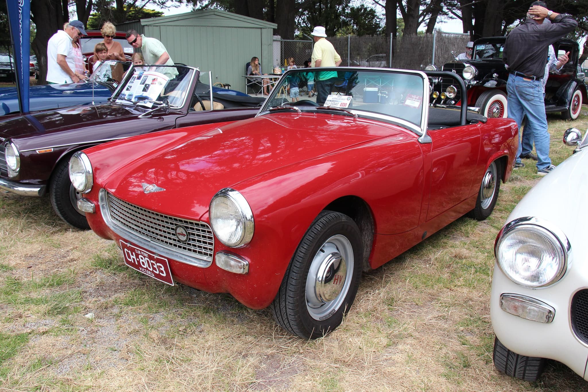 A red open two-seater with round headlamps in the wings and a mesh grille, parked on dry grass among other cars and visitors. Opens a larger view