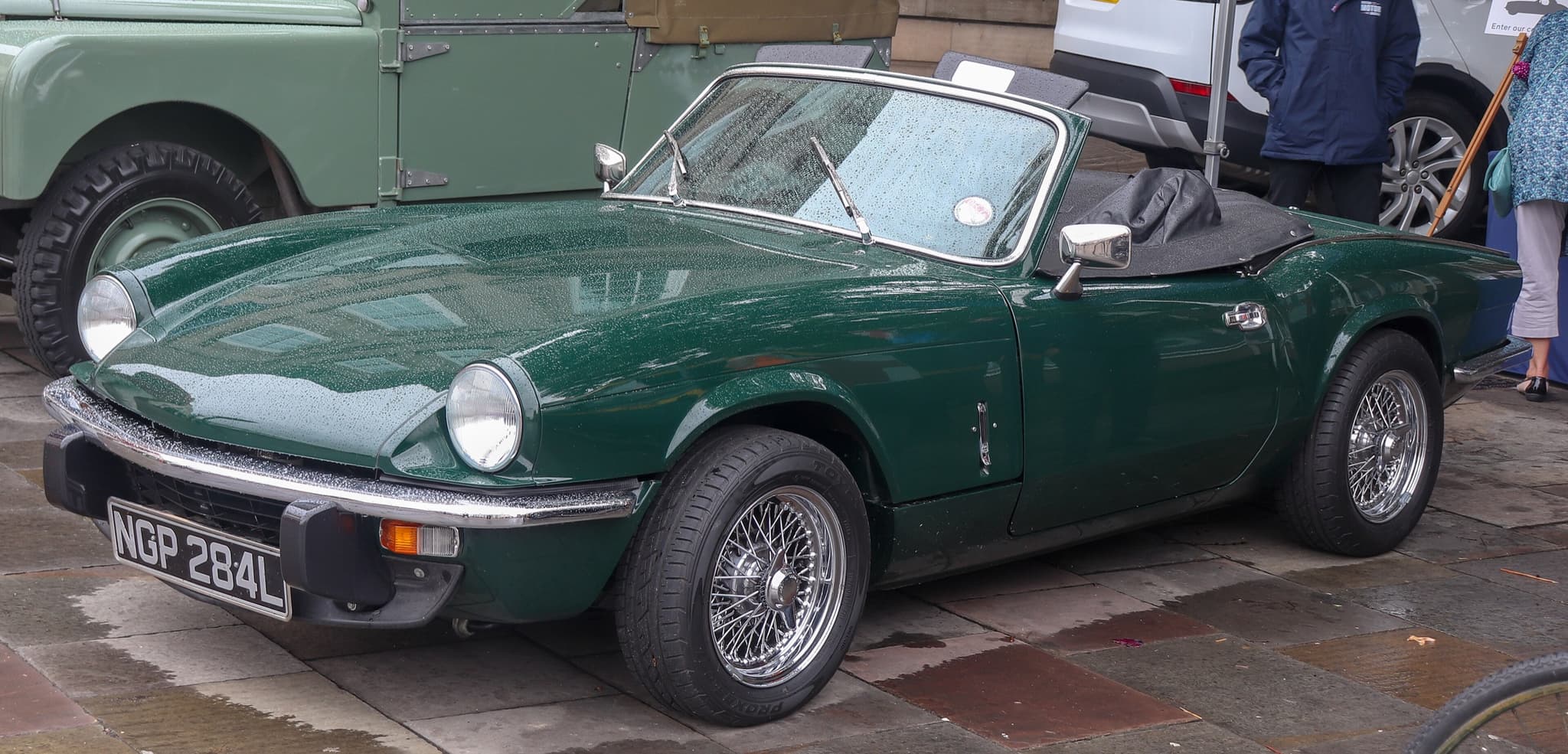 A green Spitfire with wire wheels and its hood folded, beaded with rain on wet stone paving.