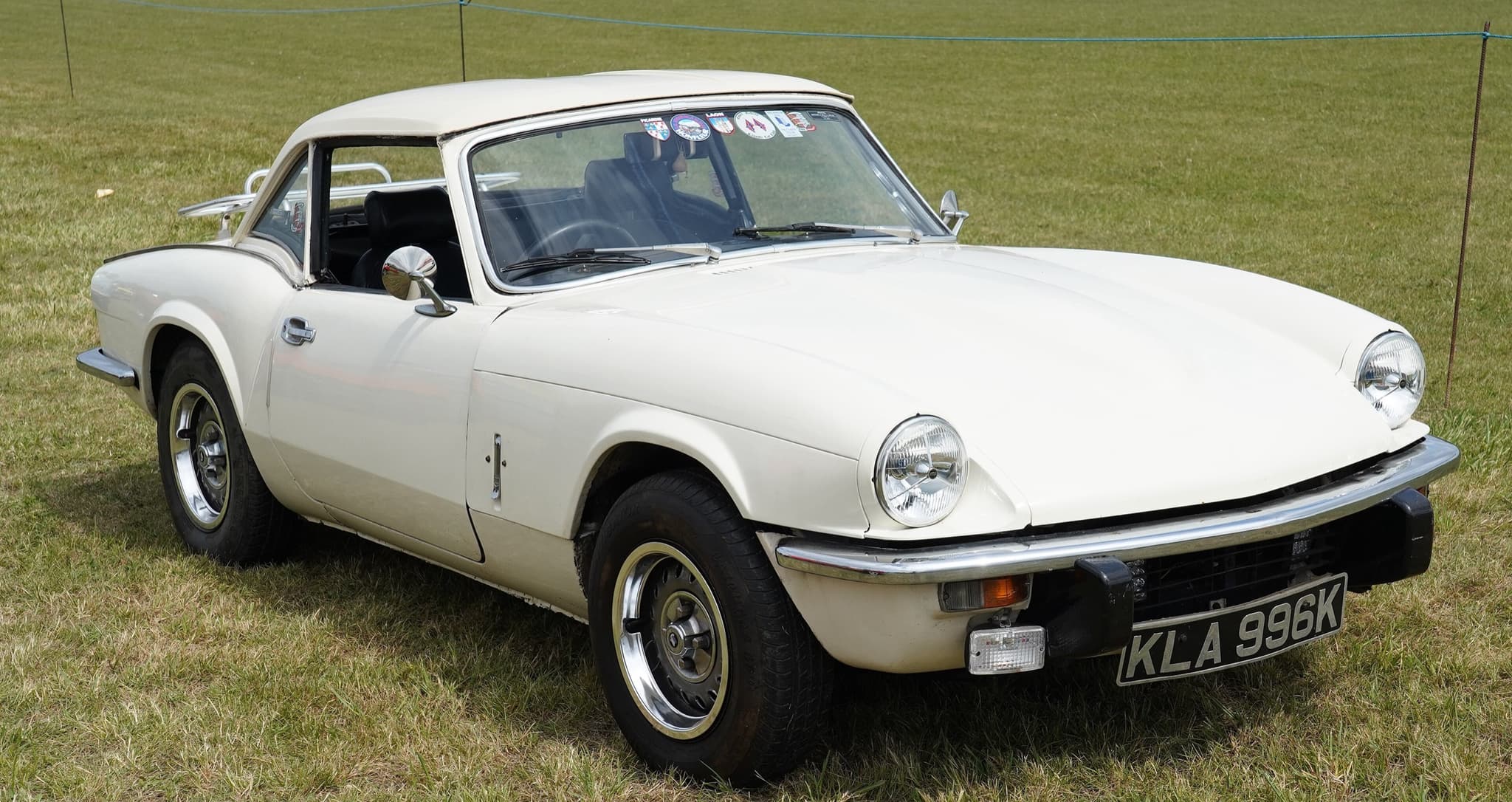A white Spitfire with a matching white hardtop on dry grass at an outdoor show. Opens a larger view