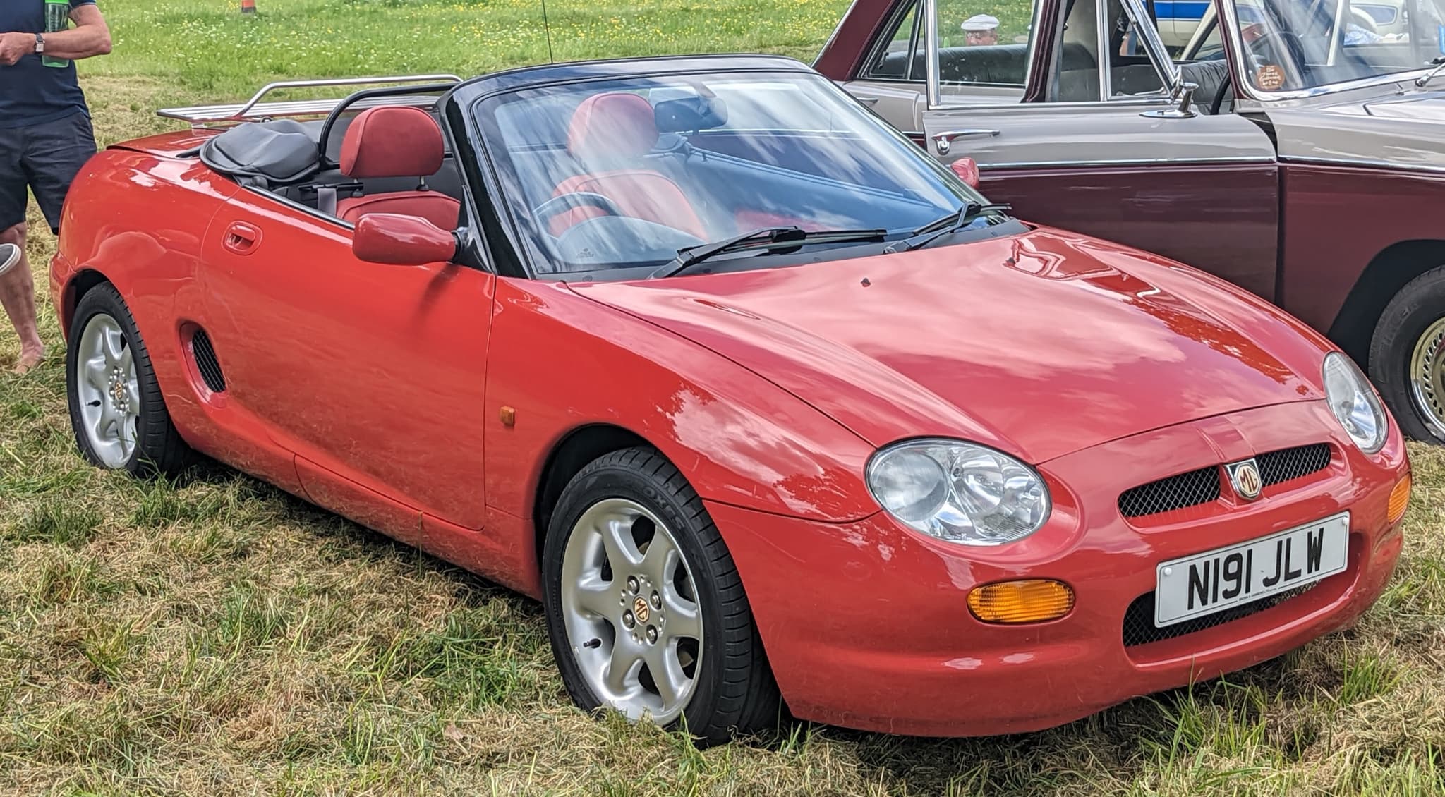 A red mid-engined roadster with its hood down and red seats, oval grille and large headlamps, parked on grass beside an old saloon.