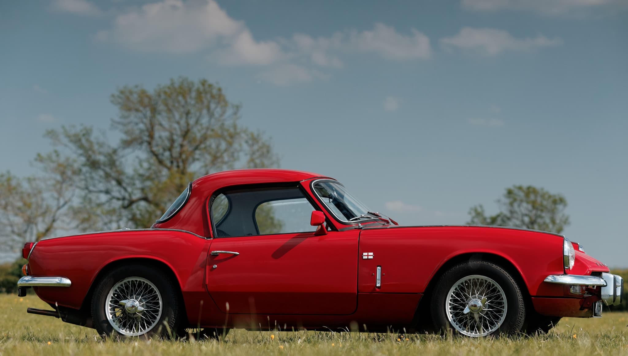 A red Spitfire Mk III with a matching hardtop and wire wheels in profile on a buttercup meadow. Opens a larger view