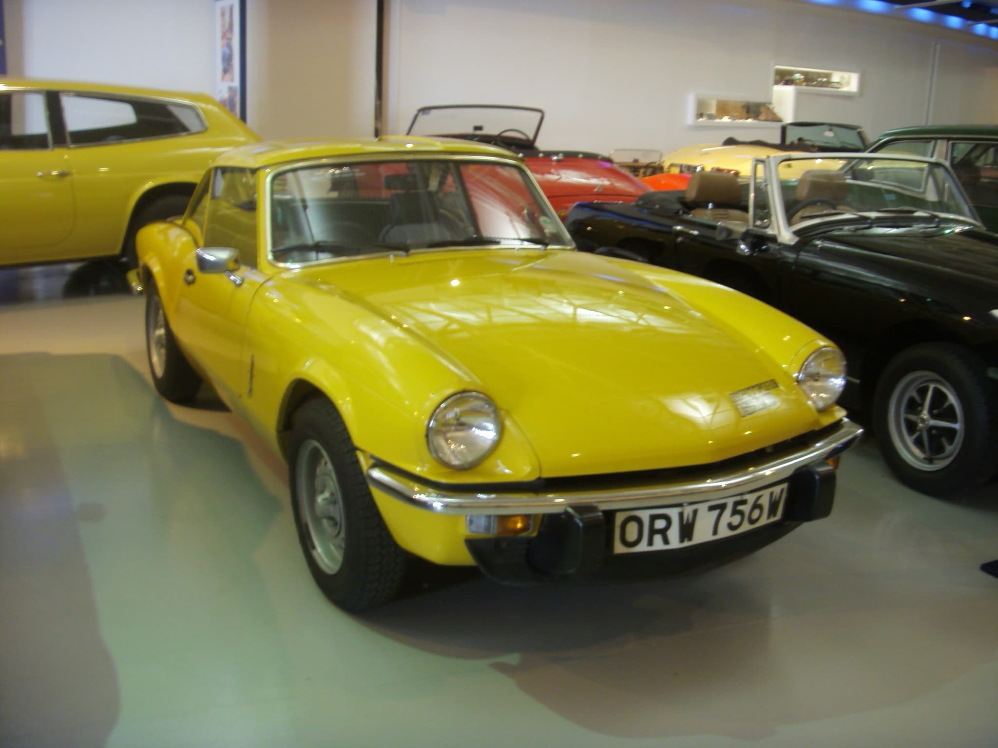 A yellow Spitfire 1500 with a matching hardtop and black over-riders on a museum floor among other cars. Opens a larger view