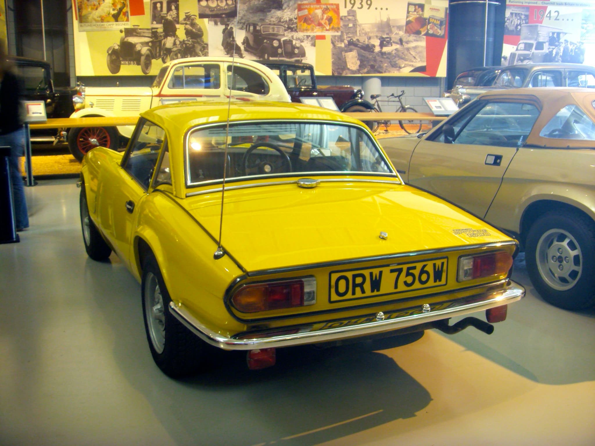 A bright yellow Spitfire 1500 with a matching hardtop, seen from the rear three-quarter in a museum hall. Opens a larger view