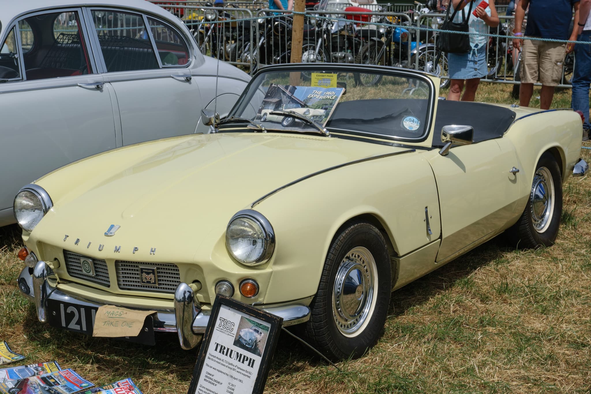 A cream Spitfire 4 with its hood stowed, on grass at a show behind a club information board. Opens a larger view