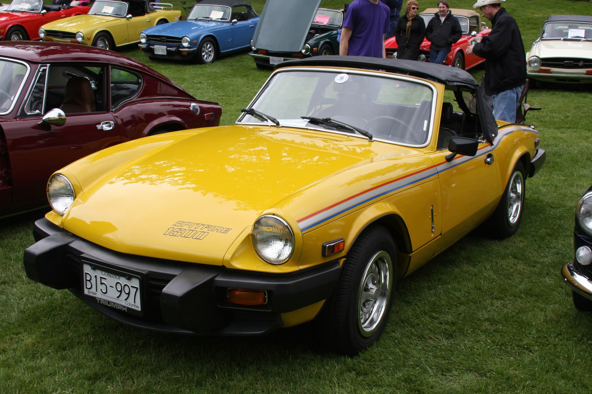A yellow Spitfire 1500 with a black rubber bumper and a red, silver and blue side stripe, on grass. Opens a larger view