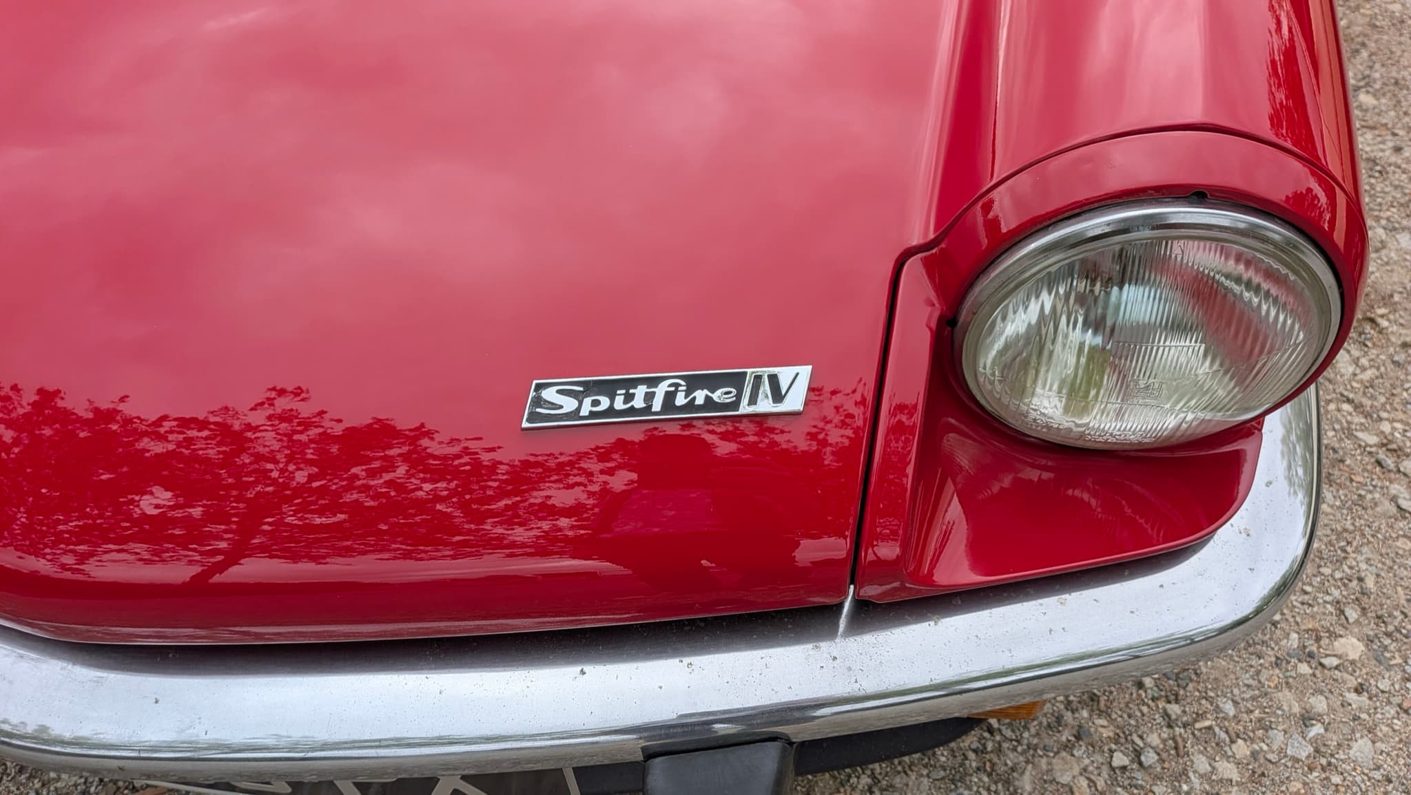 A chrome Spitfire IV badge on the red bonnet of a Spitfire, beside the headlamp and above the bumper. Opens a larger view