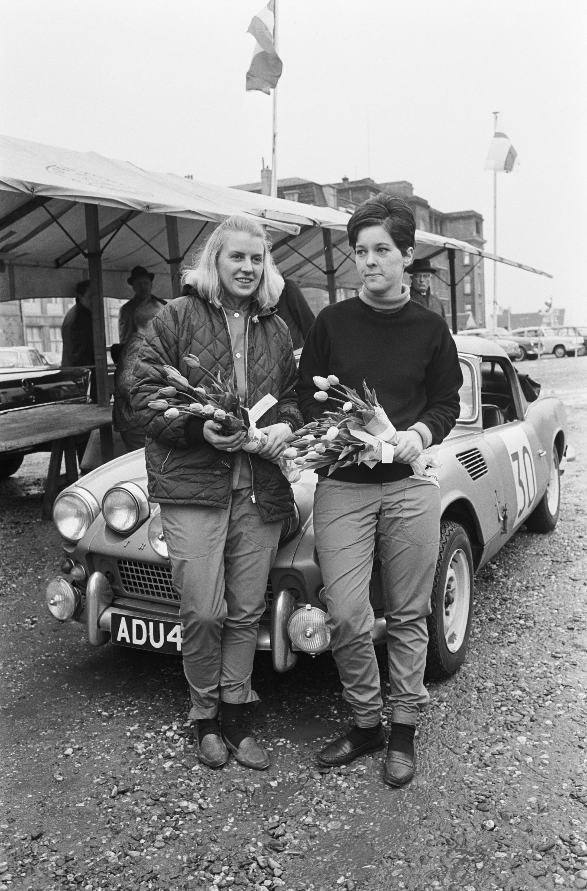 Two women in rally clothes holding tulips lean on a Spitfire 4 with auxiliary lamps and race number 30. Opens a larger view