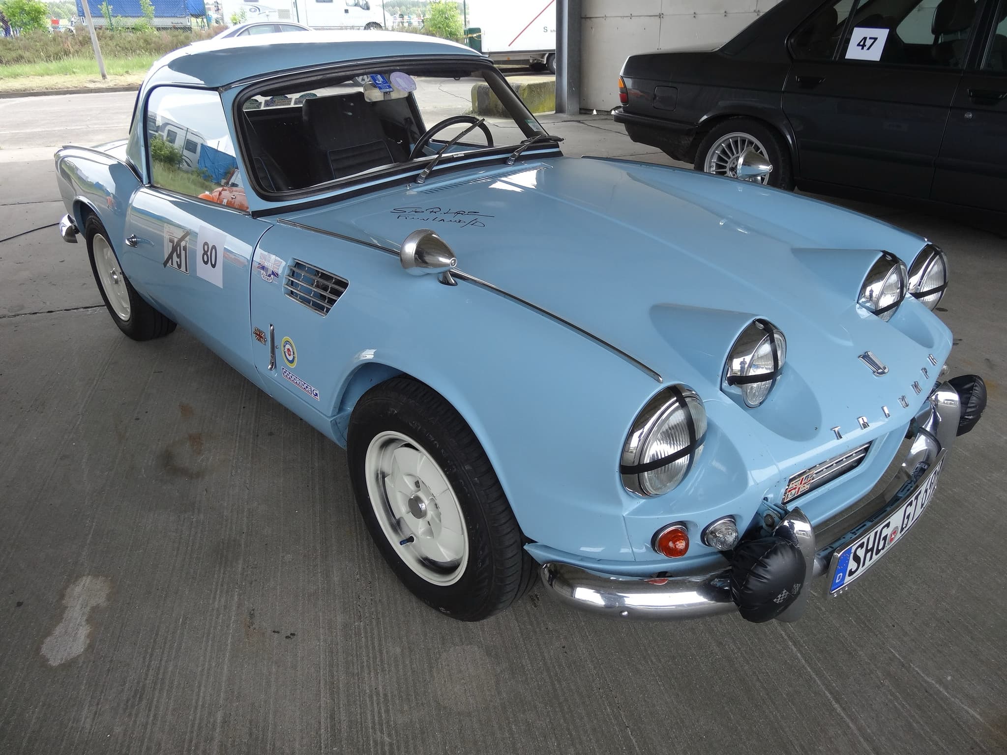 A pale blue Spitfire 4 with a hardtop, four extra lamps and door numbers, parked under a shelter. Opens a larger view