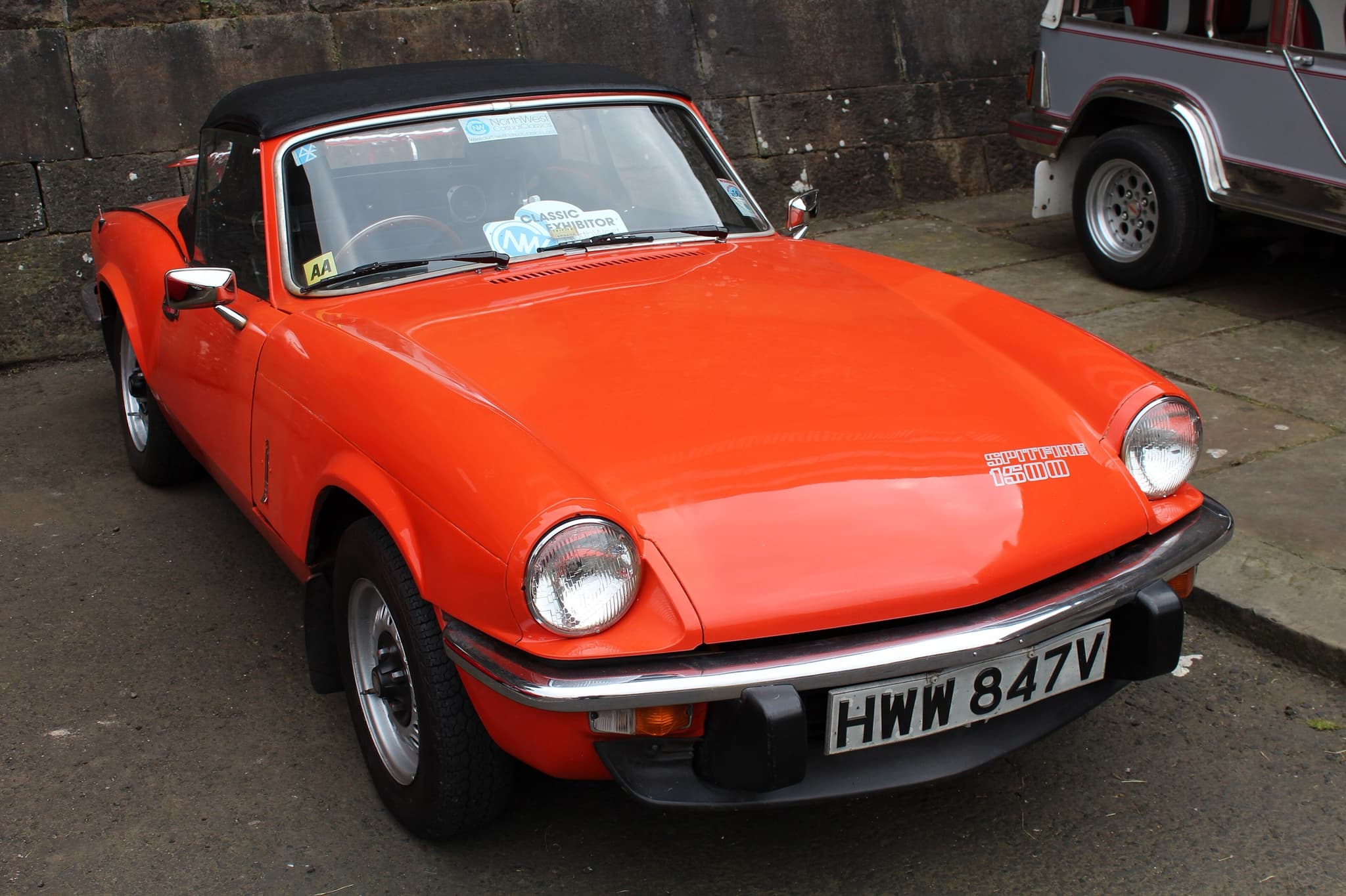 An orange Spitfire 1500 with its black hood up and a bonnet decal, parked before a sandstone wall. Opens a larger view