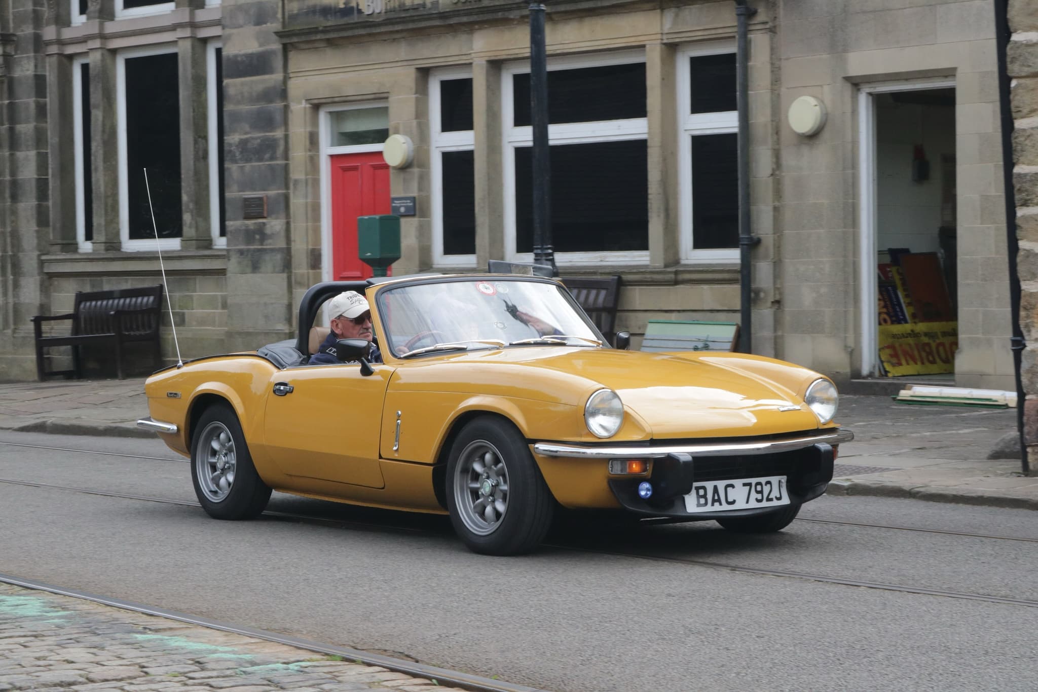 A mustard-yellow Spitfire, hood down, driven along a street with tram rails past stone buildings. Opens a larger view