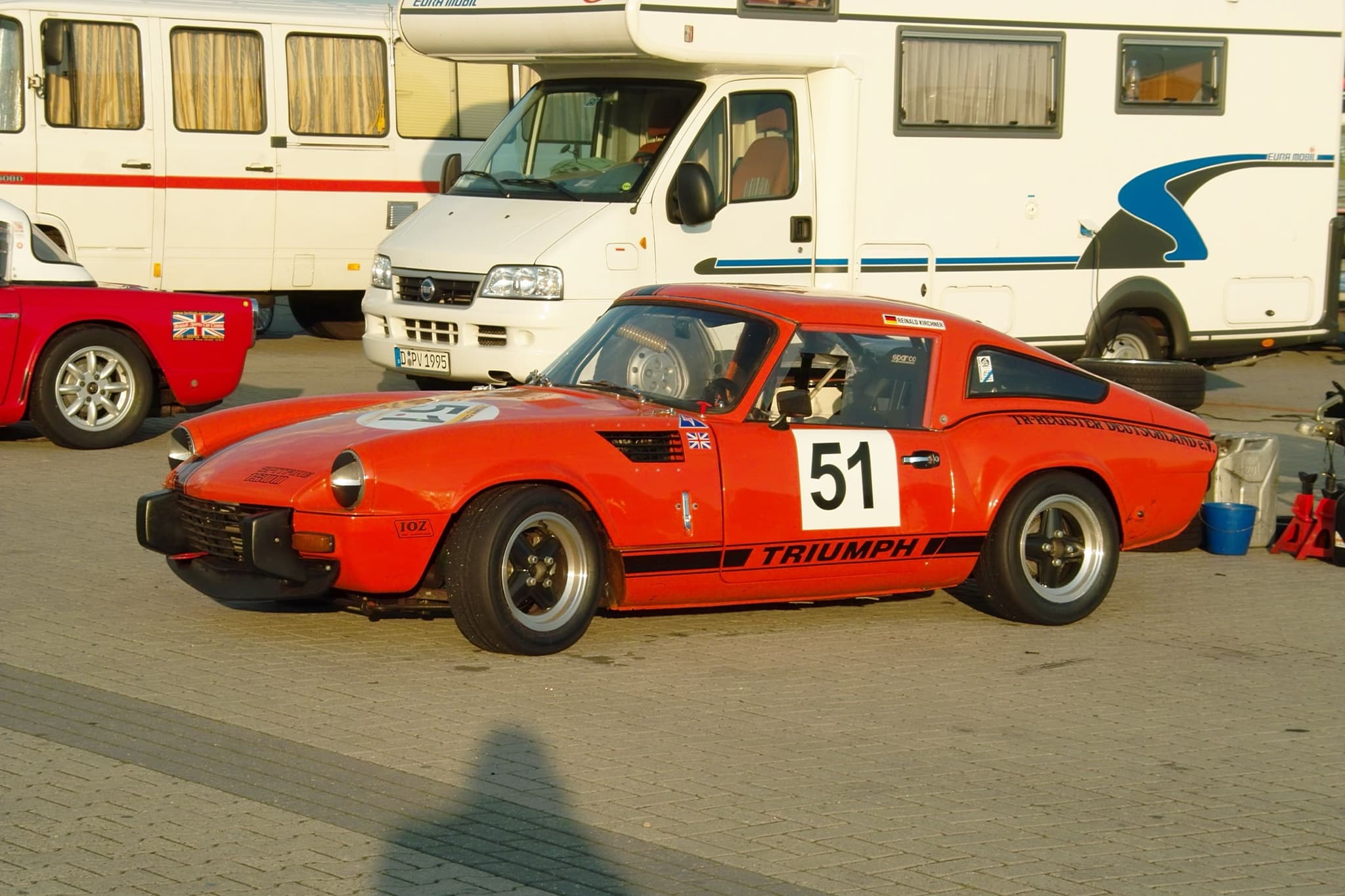An orange Spitfire race car numbered 51 with a sloping fastback hardtop, parked beside motorhomes. Opens a larger view