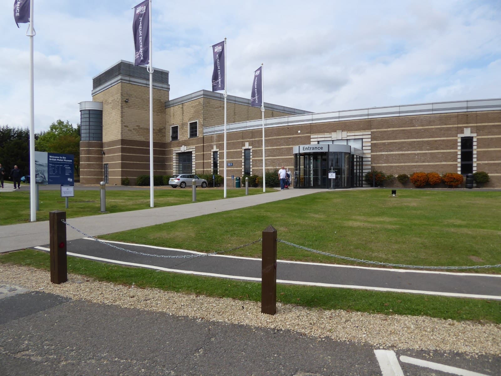 A brick museum building with a round glazed entrance, purple flags and a lawn crossed by a chain barrier.
