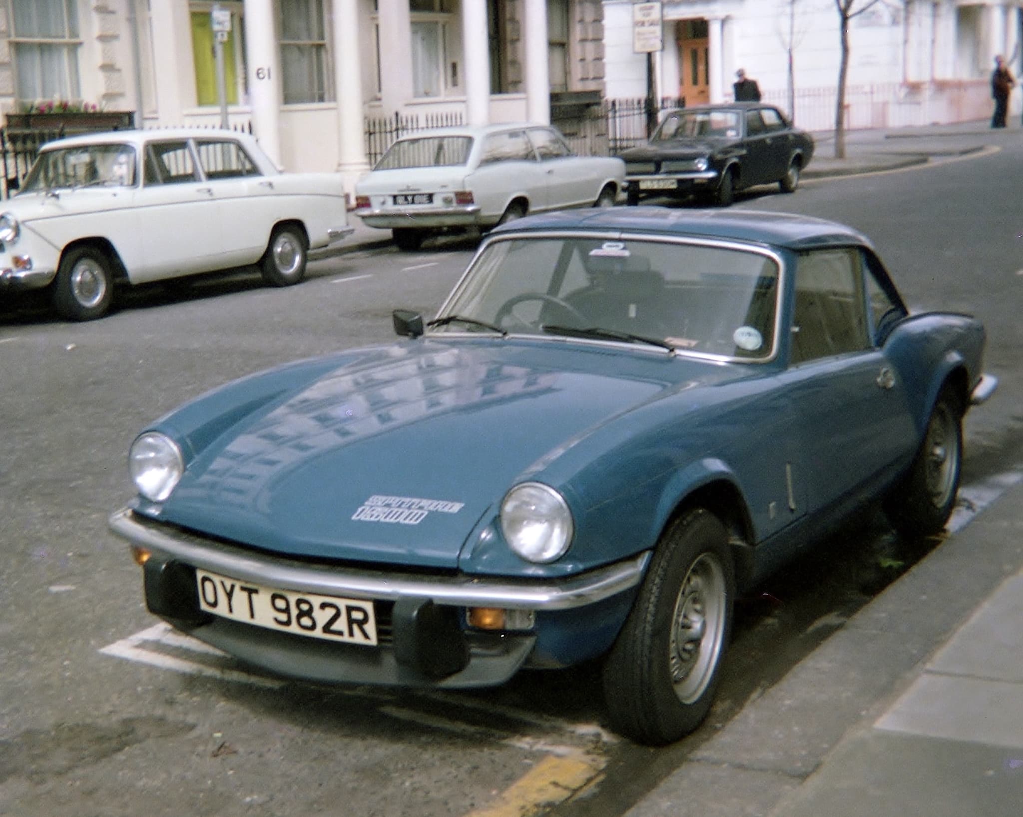 A blue Spitfire 1500 with a matching hardtop at a kerb, older saloons parked before white stucco houses. Opens a larger view