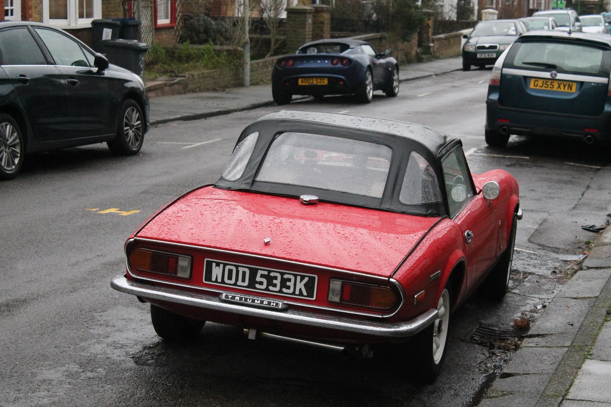 A red Spitfire in the Mk IV body with its black hood up, seen from behind on a wet street between parked cars. Opens a larger view