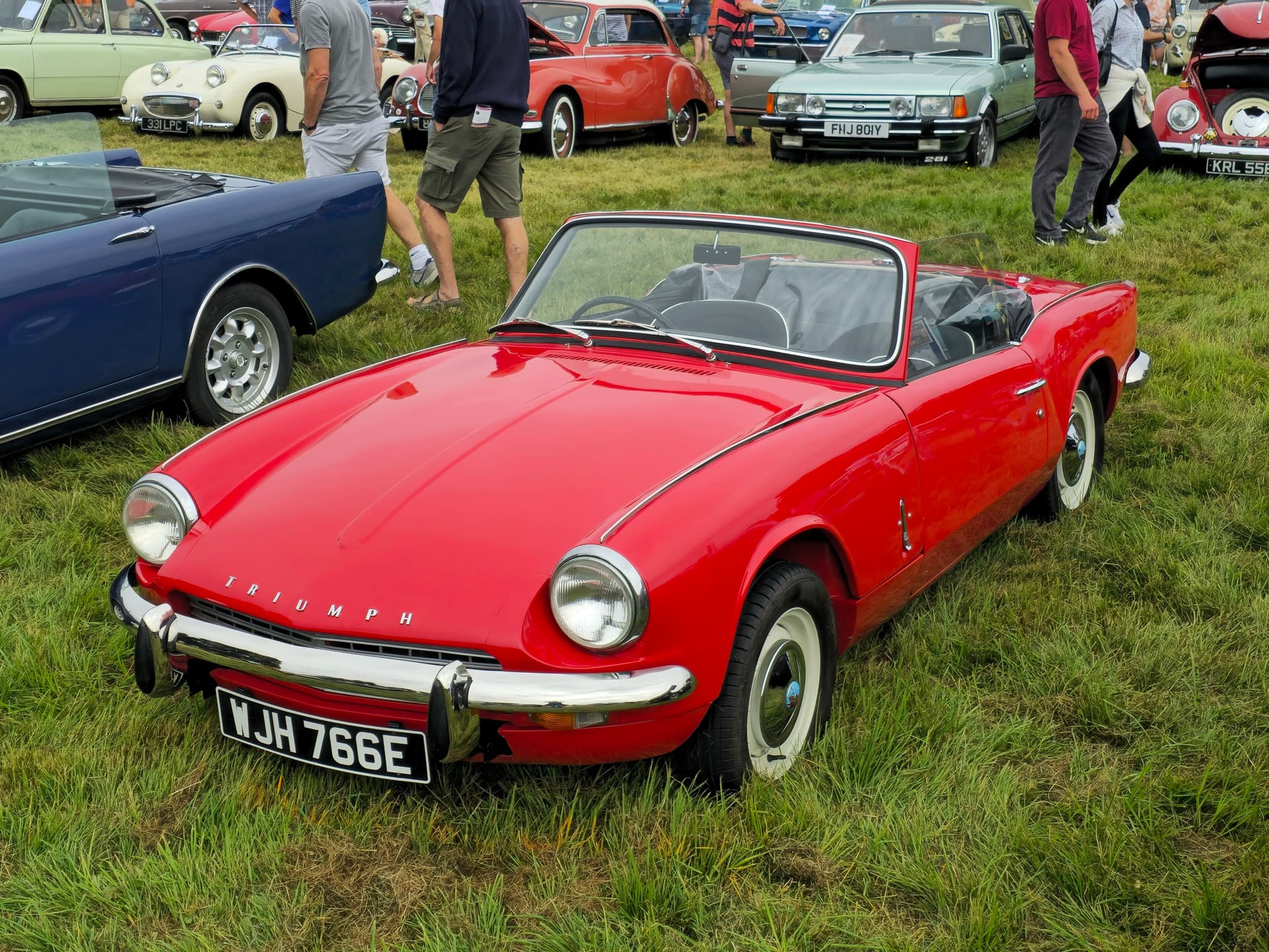 A red Spitfire Mk III with its hood folded and white-painted wheels on a show field, visitors and cars behind.