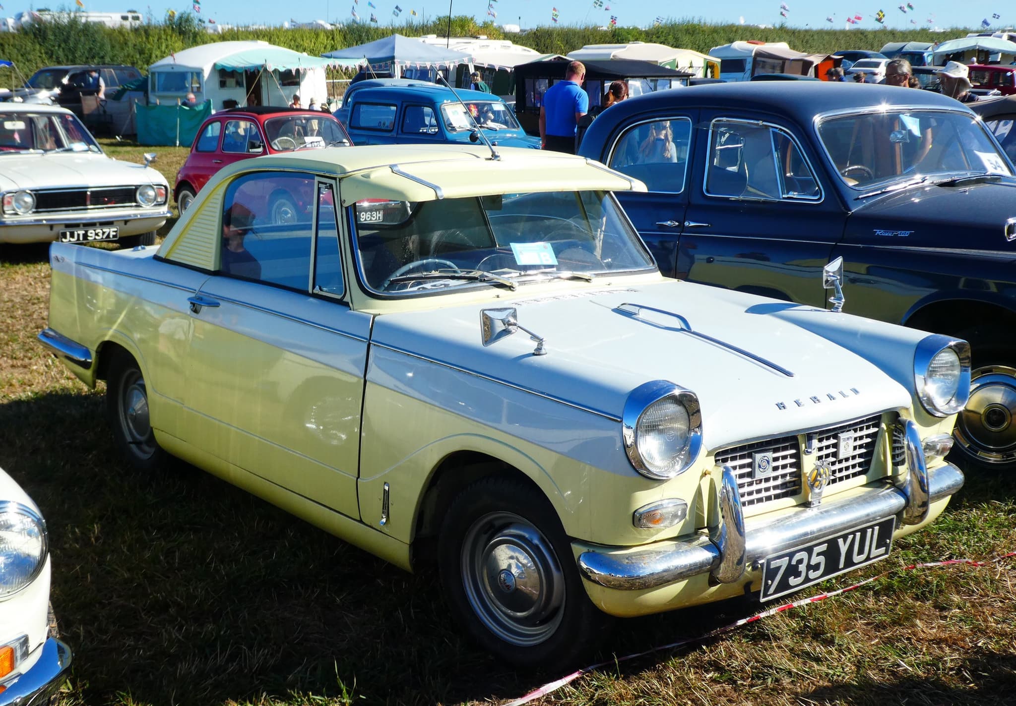 A two-tone cream and pale grey small coupé with hooded headlamps and chrome bumpers, parked on grass among other old cars at a fair.