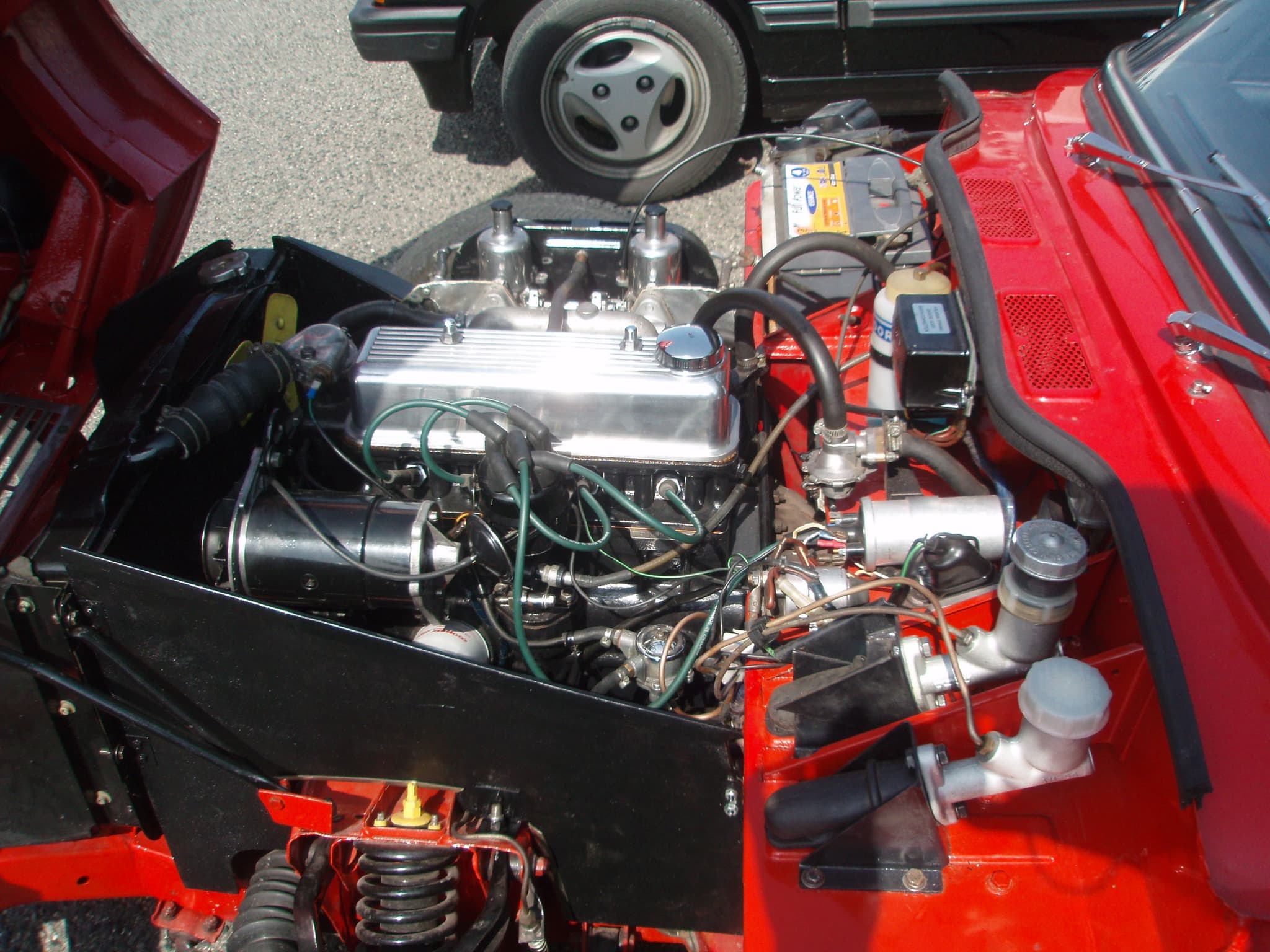 Looking down into a red Spitfire's engine bay, its bonnet tipped forward, showing twin carburettors and front suspension. Opens a larger view