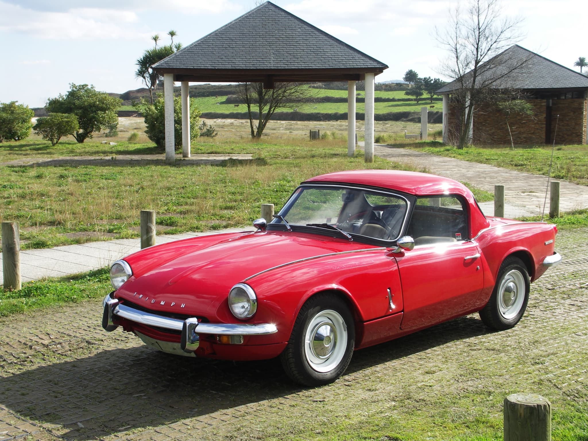 A red Spitfire Mk III with a matching hardtop and chrome bumper across the grille, parked by a park shelter. Opens a larger view