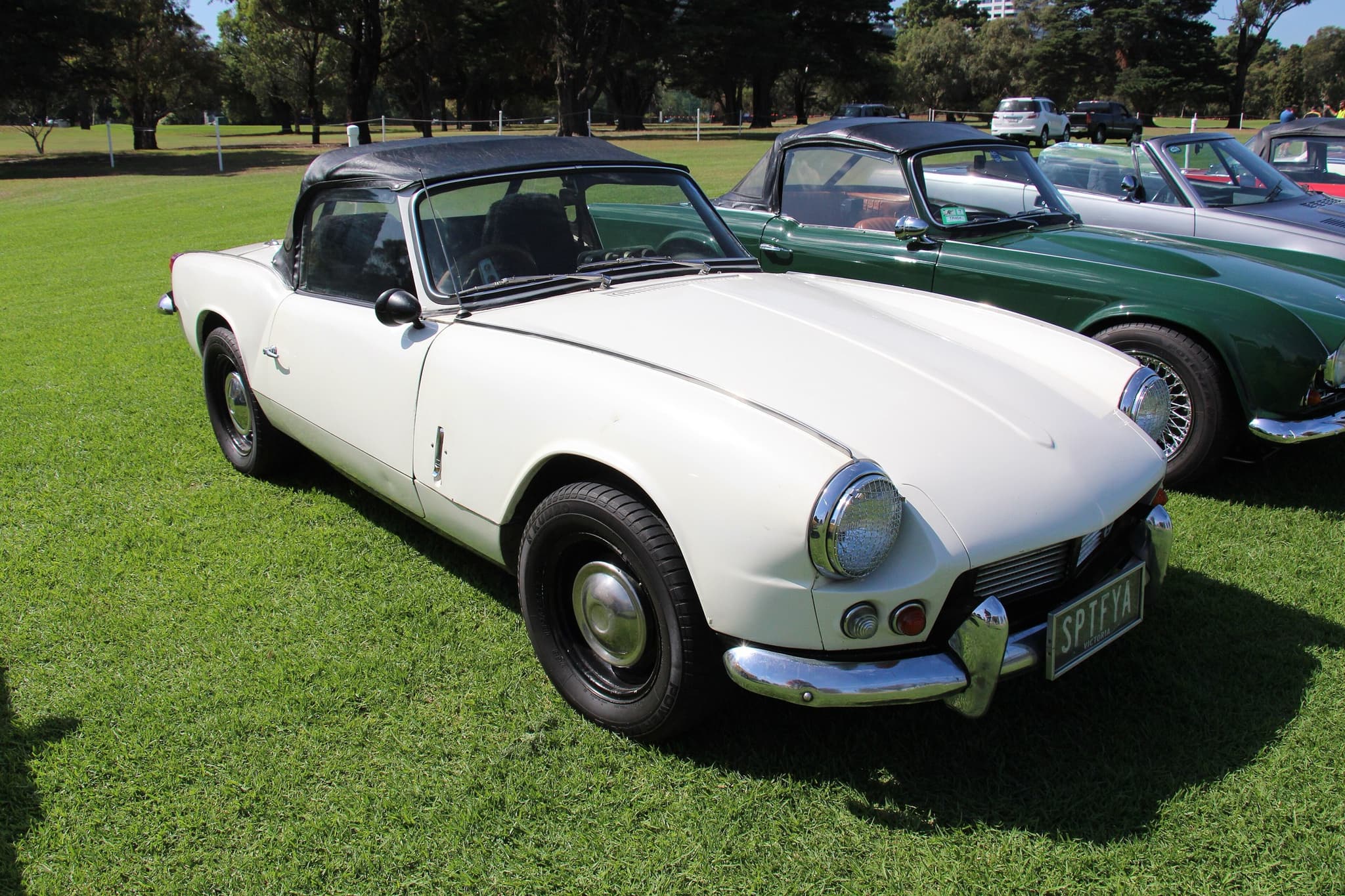 A white Spitfire 4 Mk II with a black hood up, front three-quarter on sunlit park grass beside other classics. Opens a larger view