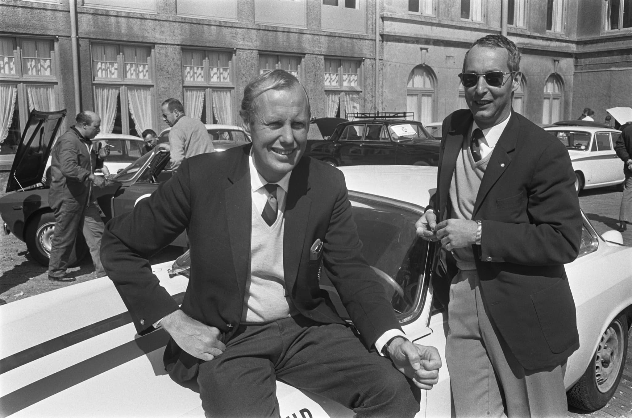 Two men in jackets and ties, one sitting on the bonnet of a white car with dark stripes, before a hotel, mechanics working behind.