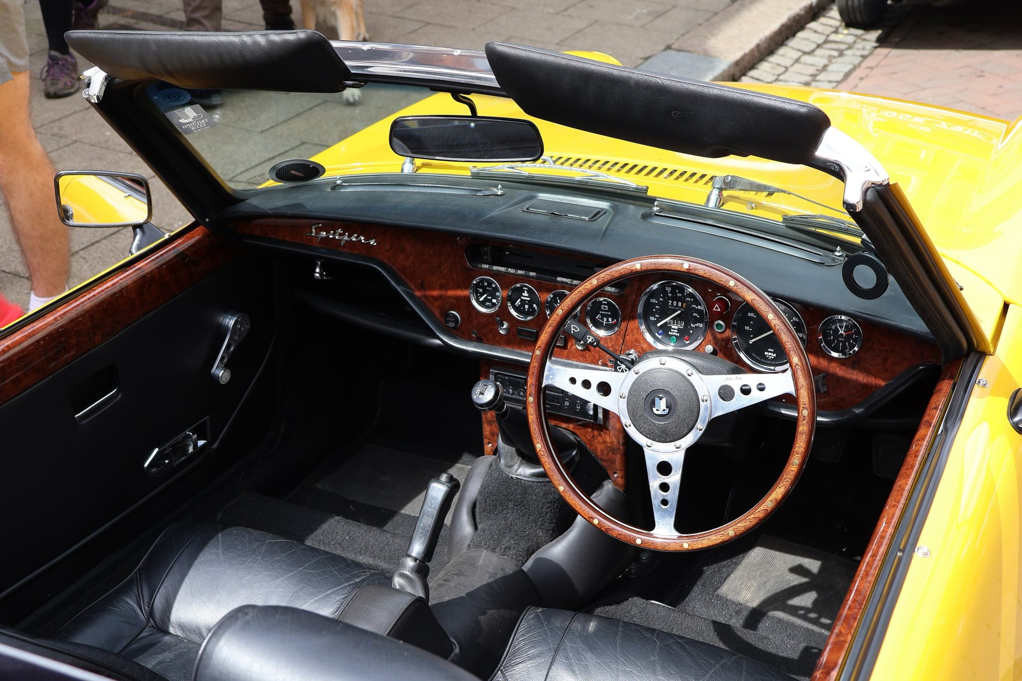 A right-hand-drive Spitfire cockpit with a burr-wood fascia, wood-rim steering wheel and black leather seats. Opens a larger view