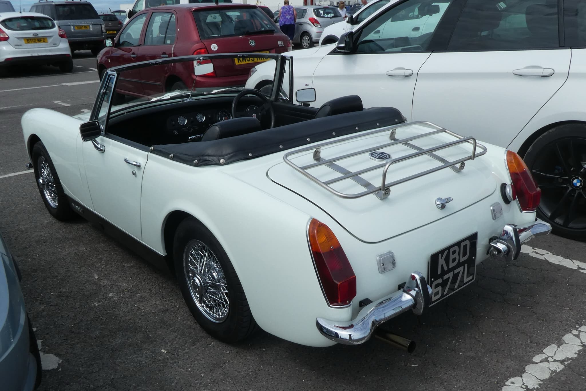 A white open two-seater from behind, with a chrome luggage rack, wire wheels and a rounded rear wheel arch, in a car park. Opens a larger view