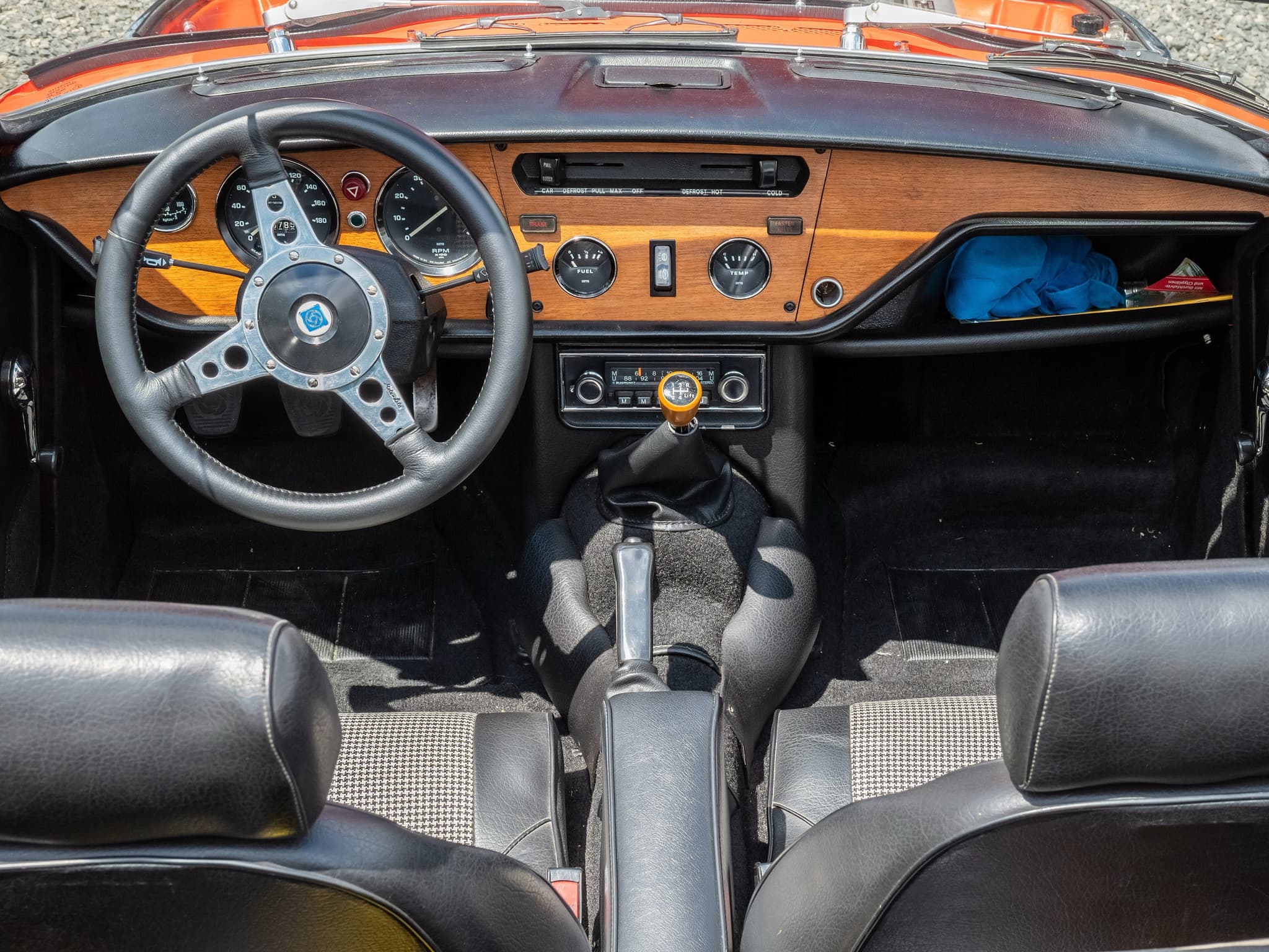 A Spitfire cockpit from behind the seats: wood fascia, leather-rimmed wheel and black seats with houndstooth inserts. Opens a larger view