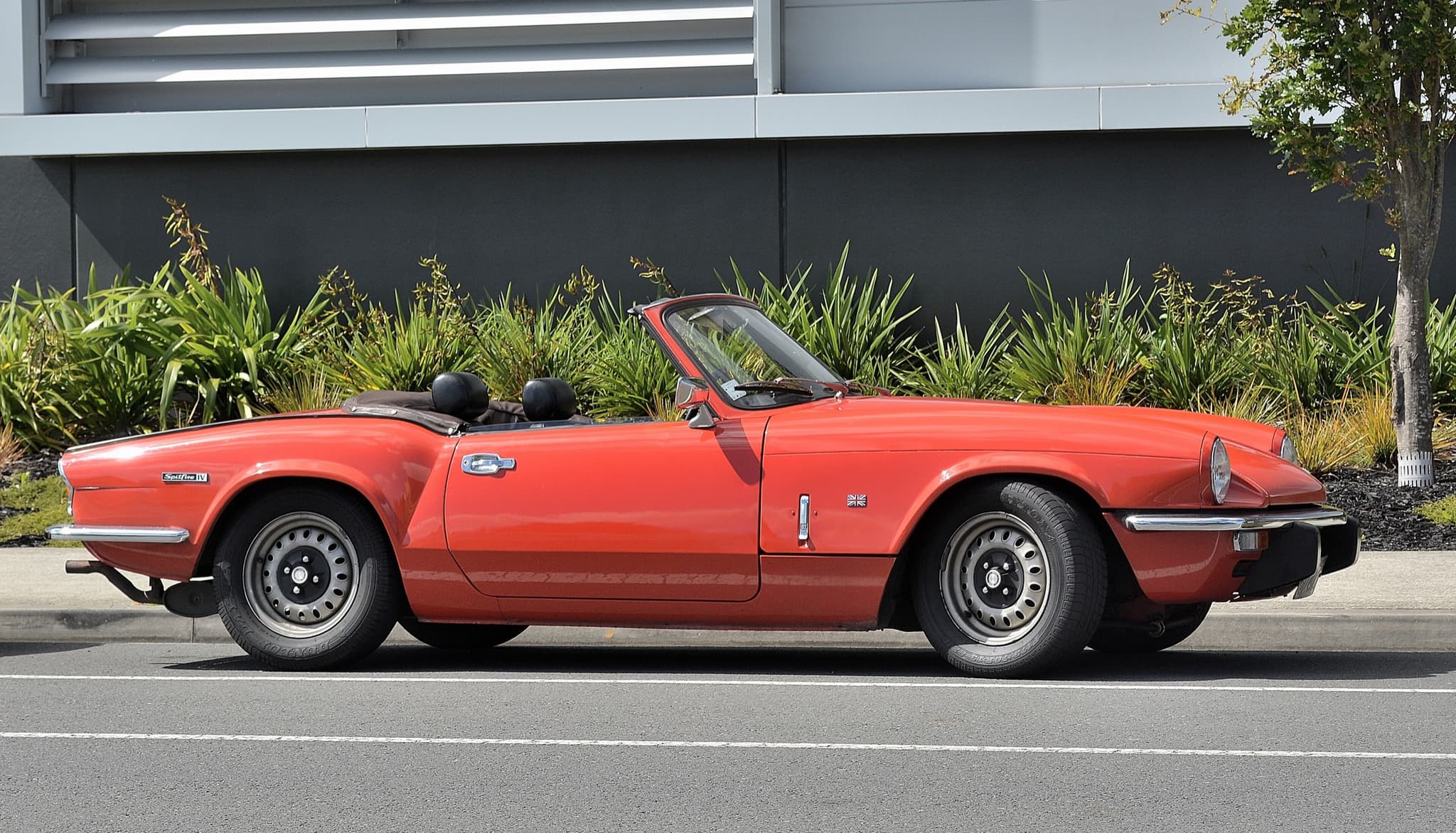 A red Spitfire Mk IV in exact side profile, hood folded, parked at a kerb before a planted bed.