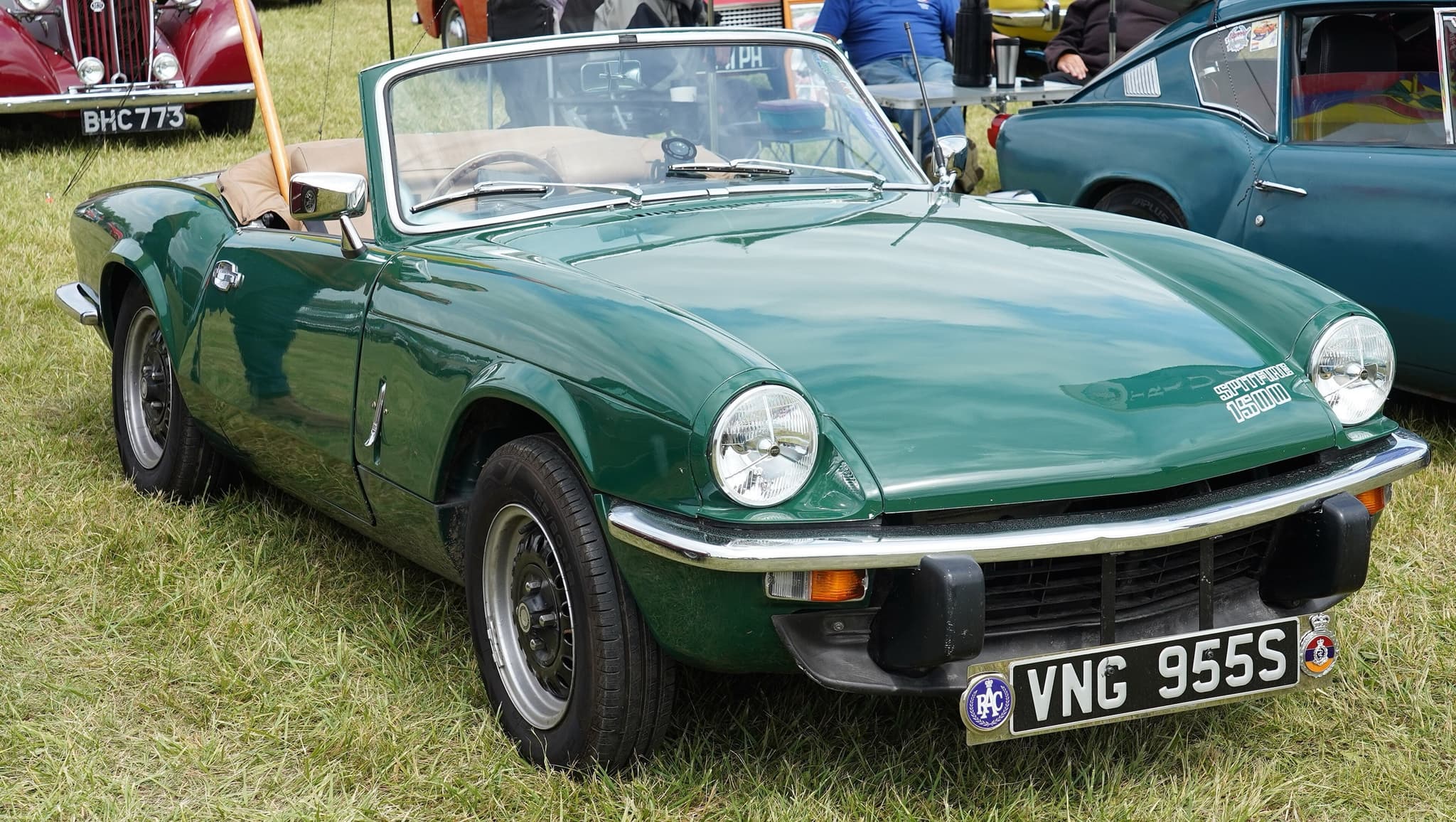 A dark green Spitfire 1500 with its hood folded and tan seats, on a grass show field among other cars. Opens a larger view