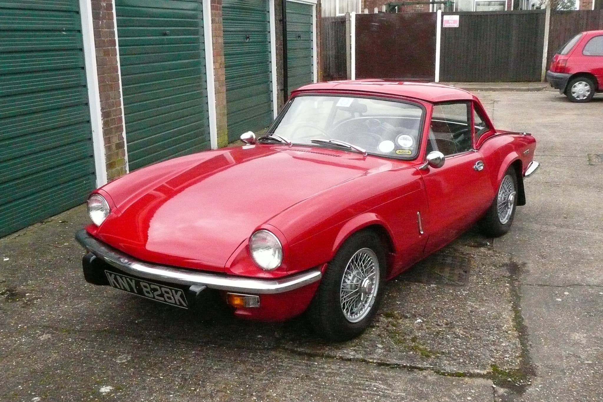 A red Spitfire with a matching hardtop and wire wheels, parked in front of lock-up garages. Opens a larger view