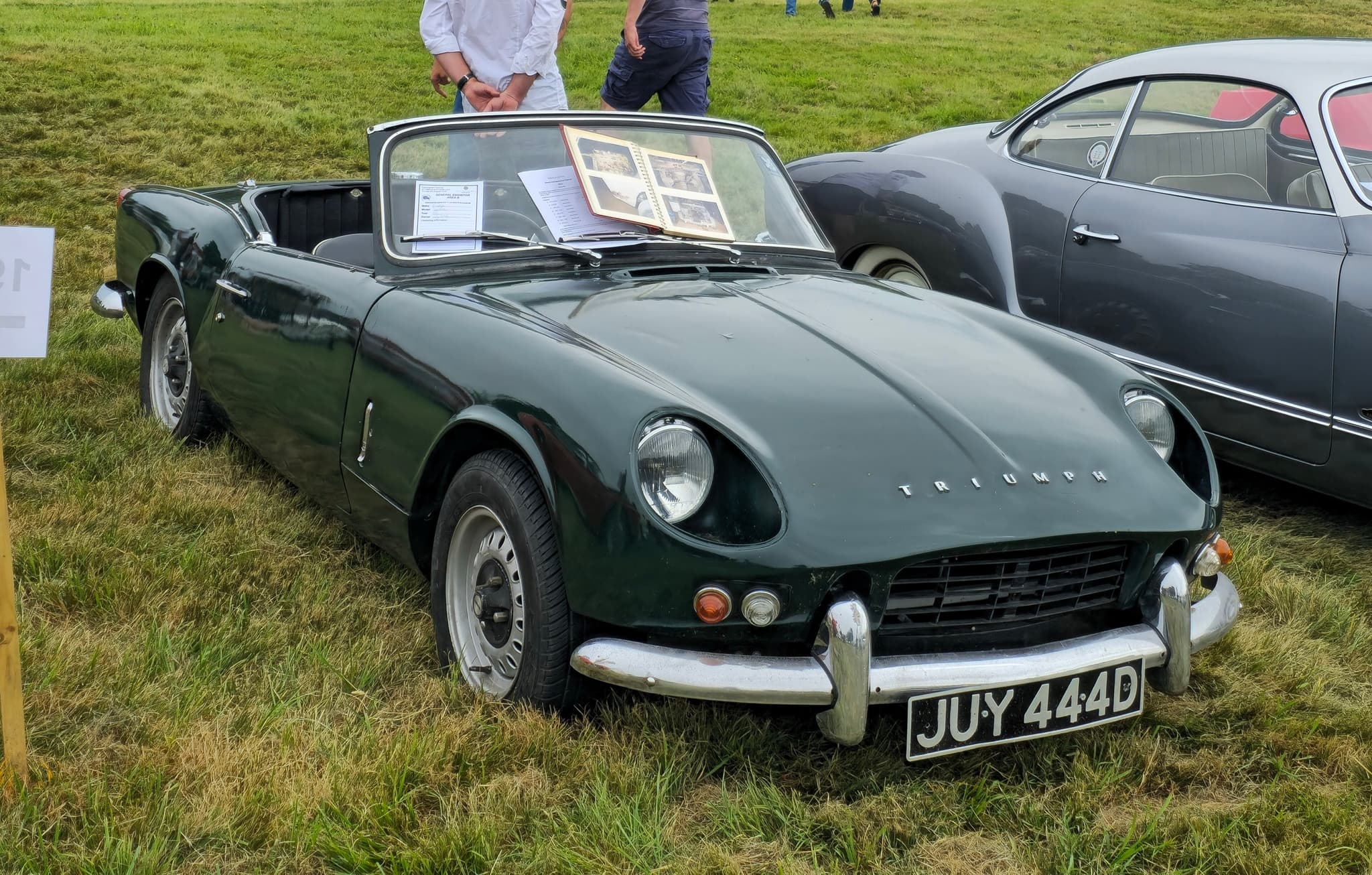 A dark green early Spitfire with its hood stowed and plain steel wheels, parked on a grassy show field.