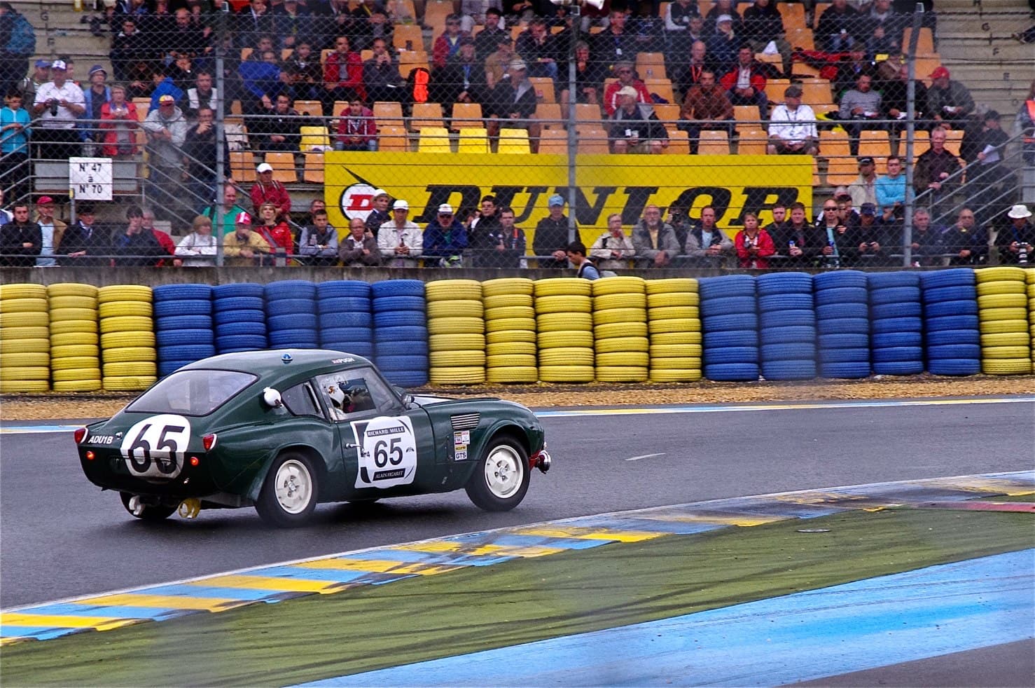 A dark green fastback Spitfire, number 65, cornering past tyre walls and a crowded grandstand. Opens a larger view