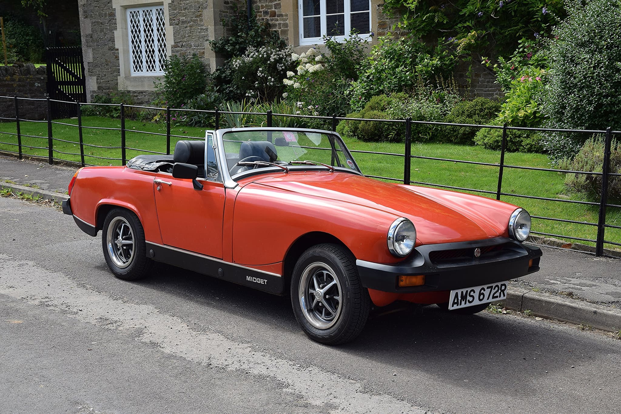 An orange open two-seater with black rubber bumpers and MIDGET lettering on the sill, parked by iron railings in front of a stone house. Opens a larger view