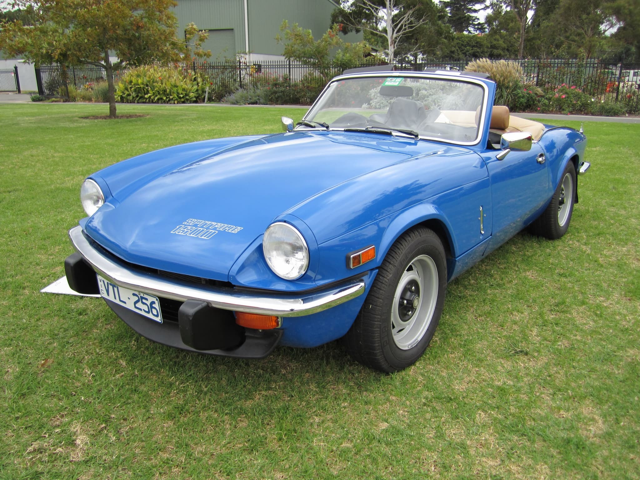 A bright blue Spitfire 1500 with tan seats and its hood folded, parked on a lawn before trees. Opens a larger view