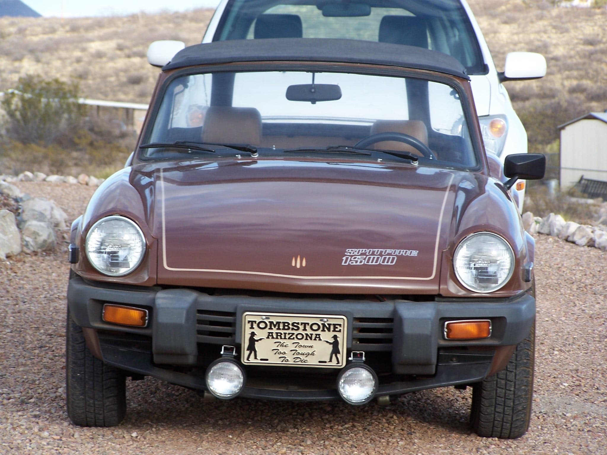A brown Spitfire 1500 with black rubber bumpers and added driving lamps, hood up, square on in the desert.