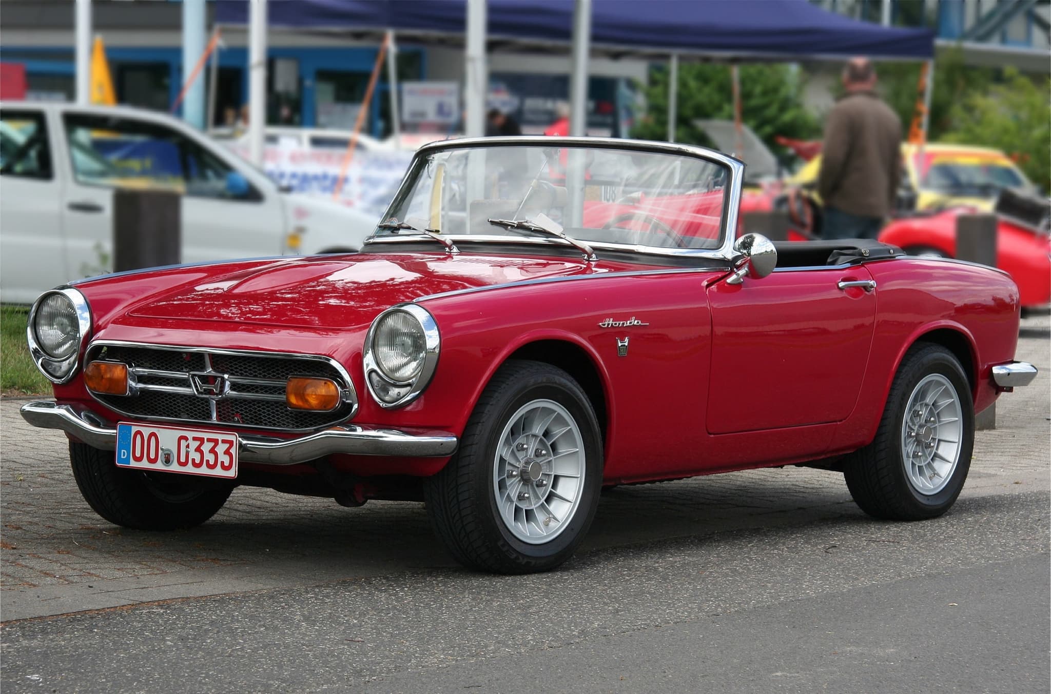 A small red roadster with chrome-ringed headlamps, a bar grille with amber lamps and a bonnet bulge, at a car show. Opens a larger view